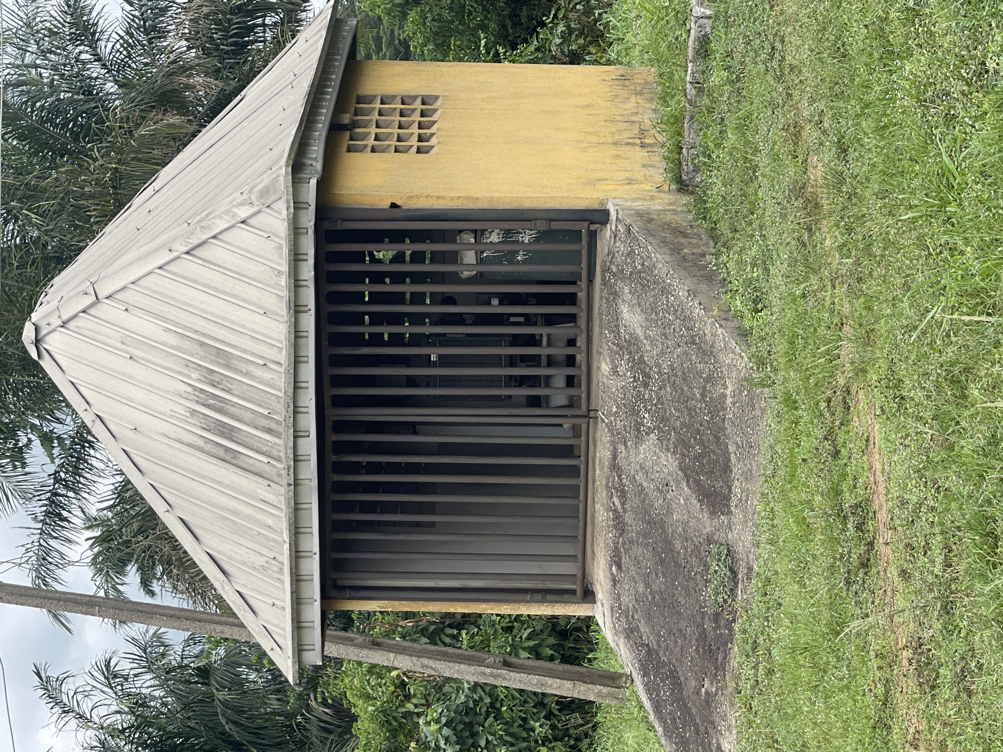 A small, yellow, concrete building with a pyramidal, metal roof sits in a grassy area, likely in a rural or suburban location. The building has a gated entrance made of metal bars. A concrete walkway leads up to the building. The building features a square latticework vent near the top. Trees and other vegetation surround the building. The location is Abraka, Nigeria.