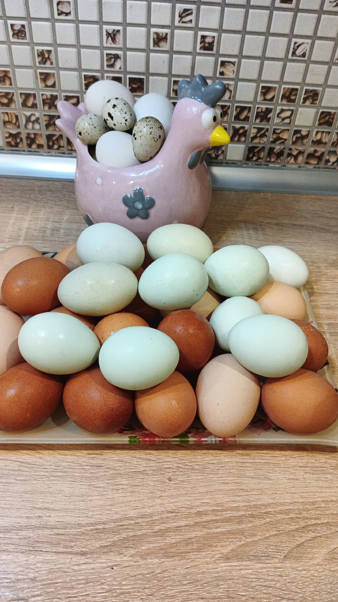The image depicts a collection of chicken eggs, some of which are speckled quail eggs, arranged on a wooden surface. In the background, a pink ceramic chicken-shaped egg holder is visible, containing more white and speckled eggs. The eggs are of various colors, including white, light blue, and brown. The overall scene appears to be set in a domestic environment, likely a kitchen, given the tiled backsplash in the background. There are no people present in the image. The image was taken in Kryvyi Rih, Ukraine.