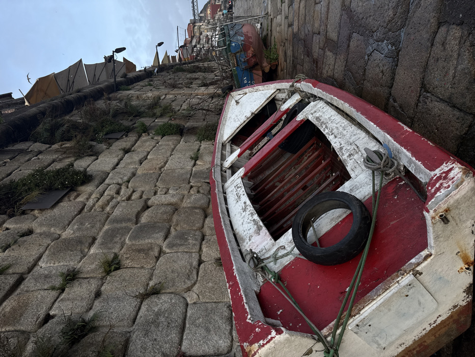 An old, red and white wooden rowboat sits askew on a cobbled walkway. The boat contains a black tire and some weathered rope. Beyond the boat, the walkway ascends a slope lined with buildings and awnings, receding into the background. The walkway itself is made of large, rectangular cobblestones, some with small patches of vegetation growing between them. A sturdy wall forms a boundary to the right of the boat. The sky is visible in the upper left corner.
