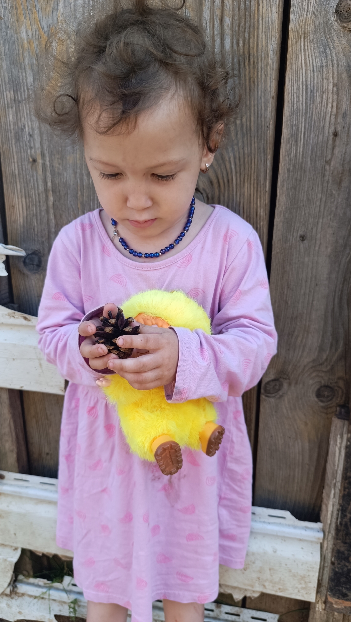 A young Caucasian child with curly brown hair stands outdoors in front of a weathered wooden fence. The child is wearing a long-sleeved pink dress with a rainbow pattern and a blue beaded necklace. They are holding a fuzzy yellow toy duck and a pinecone in their hands, looking down at them. The scene appears to be in a rural or backyard setting. No specific details about the city of Karsha, Russia, are visible in the image.