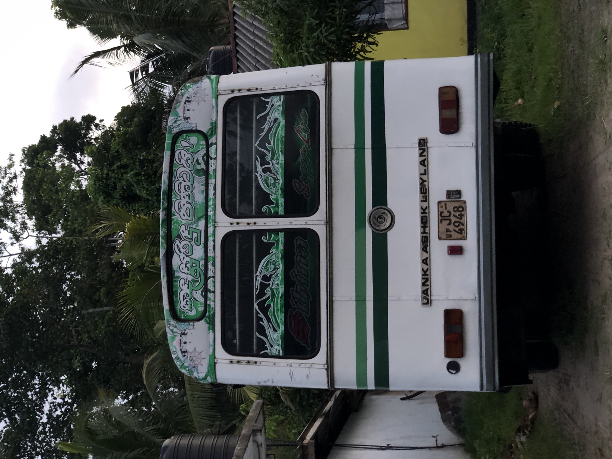 This media file captures the rear of a white bus with green stripes, parked outdoors in Kathaluwa, Sri Lanka. The bus features ornate green and white decals on its rear windows, with some text visible within these decals, appearing to be in a South Asian script. On the right side of the bus, text reads "LANKA ASHOK LEYLAND" vertically, and a license plate with the characters "UP JC 4948" is also visible. The bus is situated on a dirt path, with lush green vegetation, including palm trees, surrounding it. A yellow building is partially visible in the background. The lighting suggests it is daytime, possibly overcast or late afternoon, given the diffused light. The overall scene evokes a sense of everyday life in a rural or semi-rural Sri Lankan setting. There are no people visible in the frame. The bus appears to be stationary.