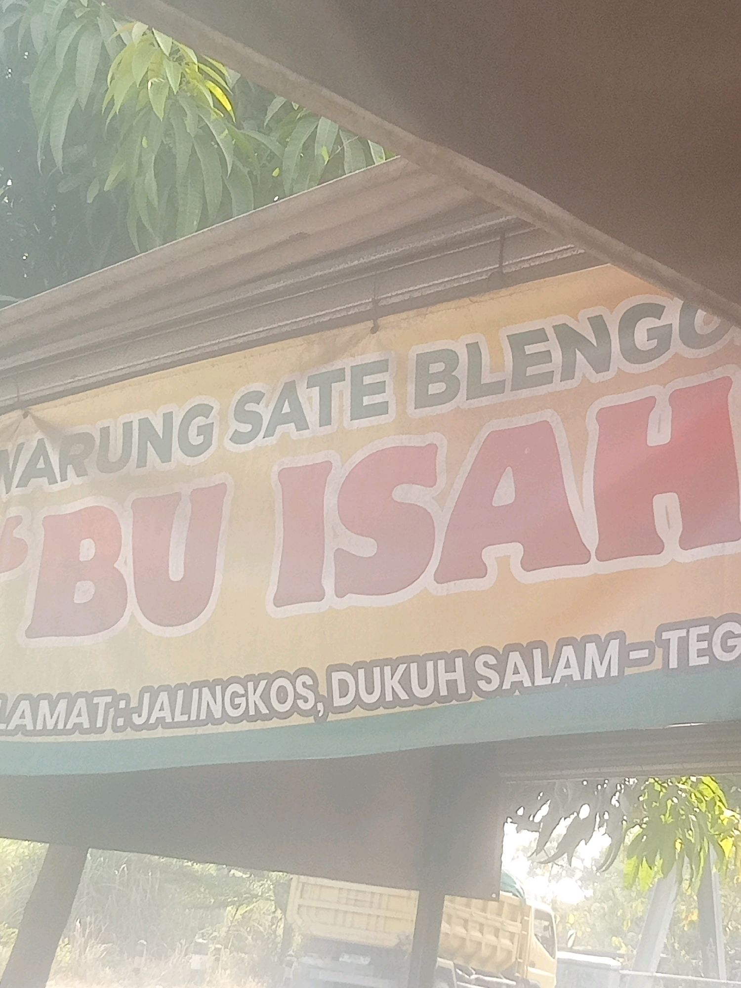 A faded banner hangs across a storefront, displaying text in Indonesian. The banner features large, pink letters spelling "BU ISAH" in the center, with "WARUNG SATE BLENGGO" above it in teal. Below "BU ISAH," the text "LAMAT: JALINGKOS, DUKUH SALAM-TEG" is visible. The banner is yellow with a green border. In the background, lush green foliage, likely trees, is visible, along with a section of a truck bed and a glimpse of a truck cabin, suggesting an outdoor setting in a semi-urban or rural area. The lighting indicates it is daytime, with sunlight filtering through the leaves. The overall impression is one of a local eatery in Dukuh Salam, Indonesia.
