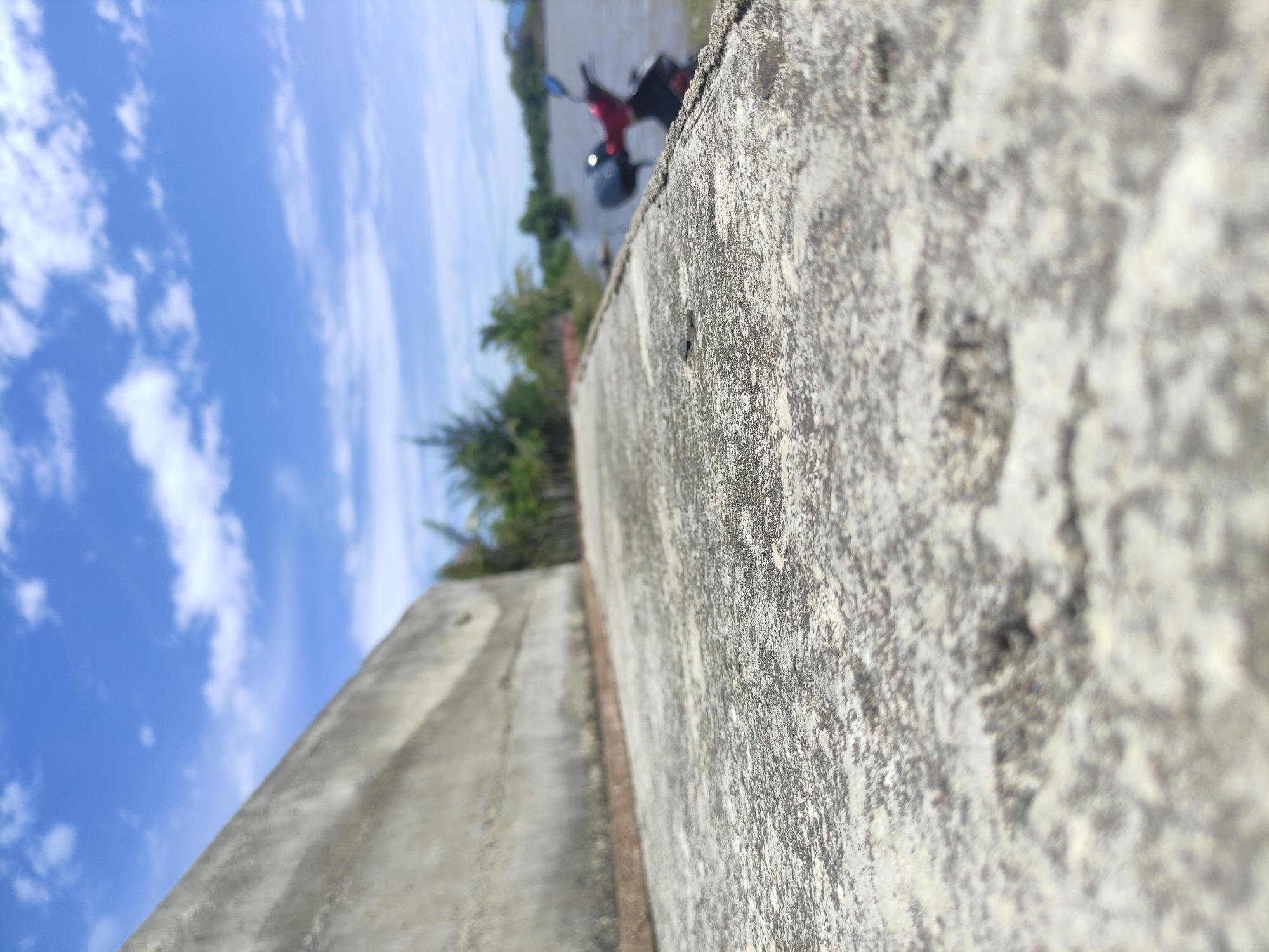 The image depicts an outdoor scene in Neuheun, Indonesia, characterized by an extreme shallow depth of field. The immediate foreground showcases a rough, textured concrete surface, heavily focused, extending from the bottom of the frame. This surface is bordered on the left by a concrete retaining wall, appearing aged and discolored, running diagonally upwards from the lower-mid-left to the mid-right. A thin, rusty-colored strip runs along the base of this wall where it meets the foreground concrete. Beyond the retaining wall and path, a body of murky water is visible. A partially obscured, out-of-focus red and black motorcycle is positioned at the water's edge on the right side of the frame. The background consists of out-of-focus green foliage, including palm trees, lining the distant bank of the waterway. Above the vegetation, a bright blue sky with scattered white, wispy clouds fills the upper portion of the image. No individuals are present, and no specific actions are observed.