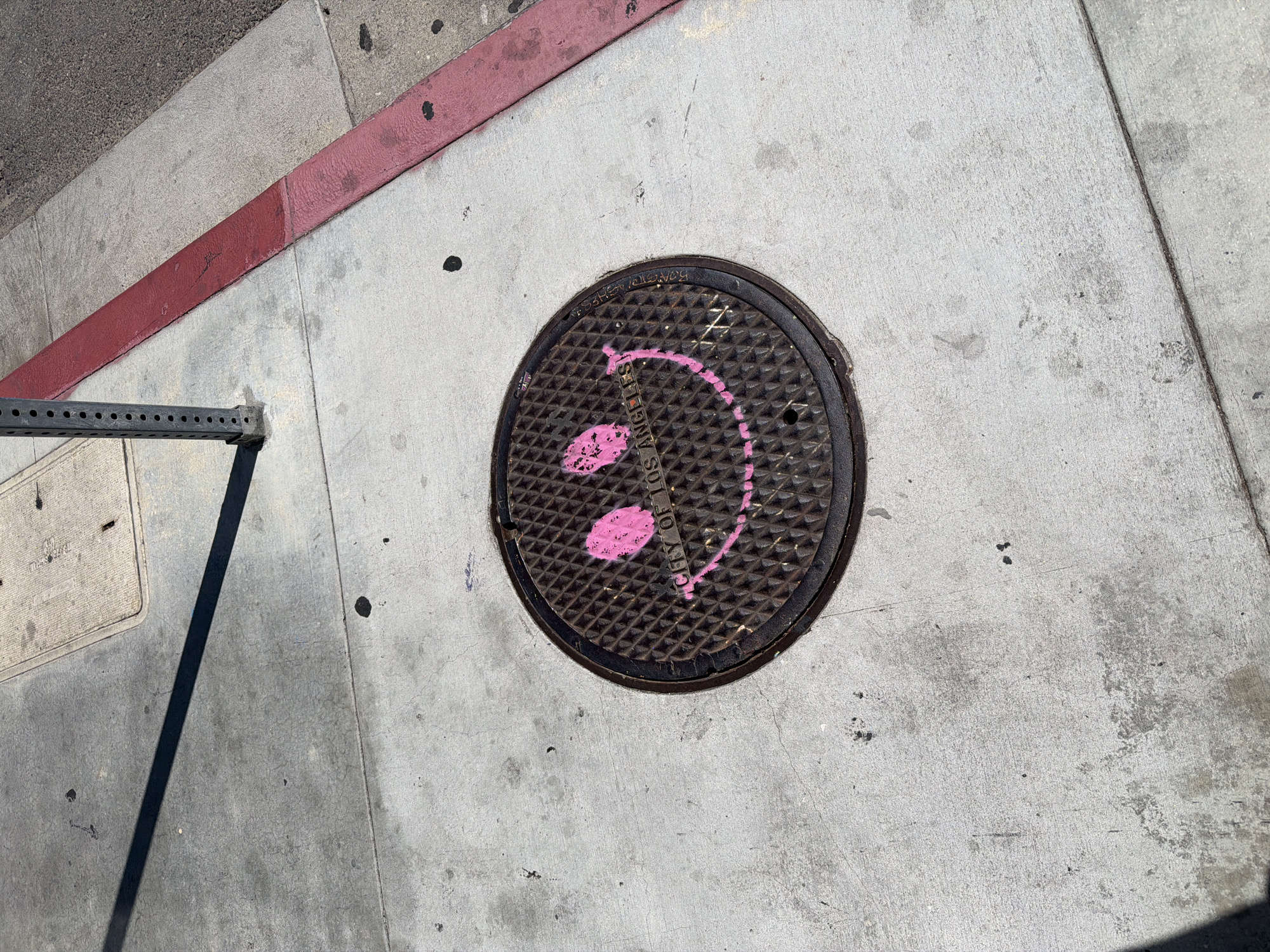 A top-down, high-angle shot captures a city street scene in Los Angeles, USA. The focal point is a circular manhole cover, painted with a whimsical pink design resembling a face, complete with eyes and a smile. The manhole cover is surrounded by a grey concrete sidewalk. A red-painted curb runs along the top left of the frame, adjacent to an area of asphalt. A metal object is positioned on the bottom left.