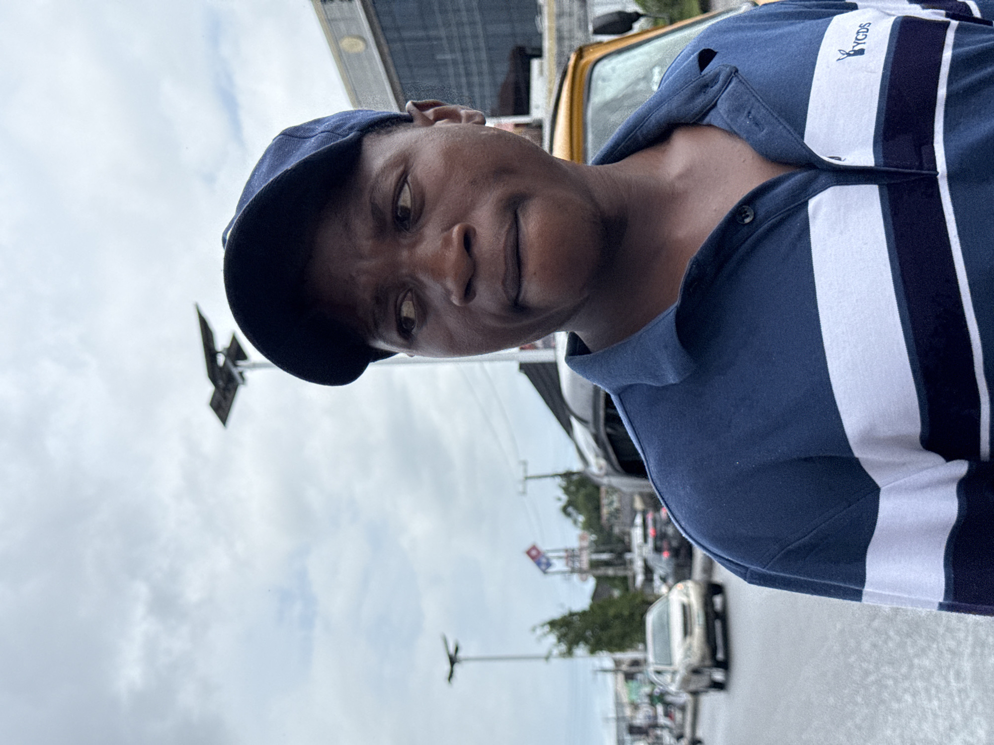 A medium, eye-level shot presents a dark-skinned person wearing a dark blue baseball cap and a blue polo shirt with thick white vertical stripes and a "YGDS" logo on the right side of the chest. The individual's face is turned slightly to the right, gazing towards the viewer with a calm expression.
The background shows a city street scene in Ado, Nigeria, with a busy thoroughfare lined with cars, vans, and overhead traffic lights. In the distance, the Domino's Pizza sign is recognizable. The sky is overcast. The scene suggests an ordinary urban environment, portraying everyday life.