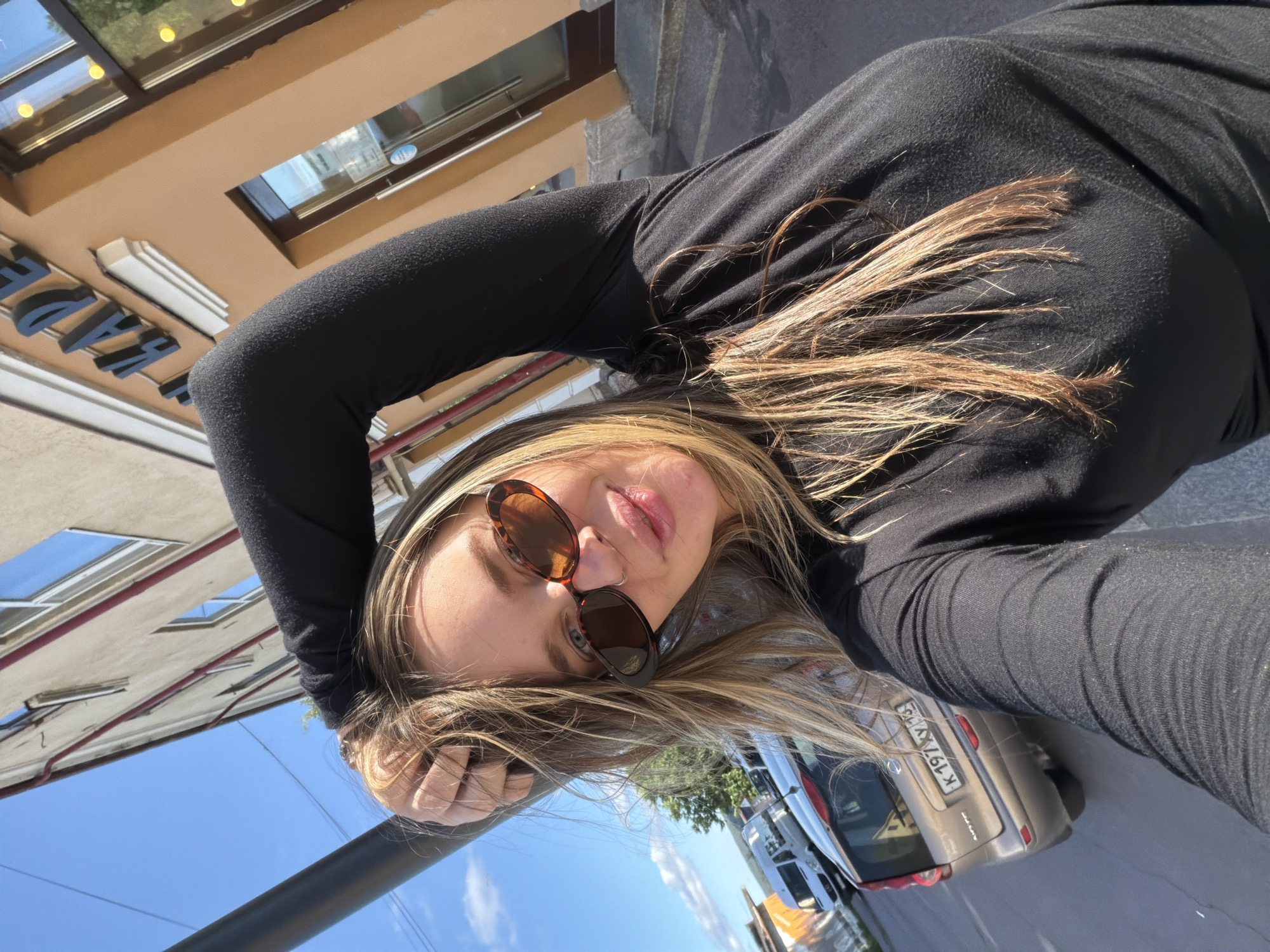 A young woman with long, blonde hair is captured in a selfie taken outdoors. She is wearing a black long-sleeved top and sunglasses with oval, tortoiseshell frames. Her left arm is raised and bent, with her hand resting on her head or neck, and her right hand appears to be supporting her head or hair. She is looking directly at the camera with a relaxed expression.
In the background, a building with beige siding and rectangular windows is visible. The sky is a clear, bright blue, suggesting it is daytime and the weather is sunny. A dark pole or railing runs vertically through the foreground. To the right of the woman, a light-colored car is parked, with its license plate visible, reading "K 197YX 123". The scene appears to be a street or outdoor public area in Sortavala, Russia. The overall mood is casual and sun-drenched.