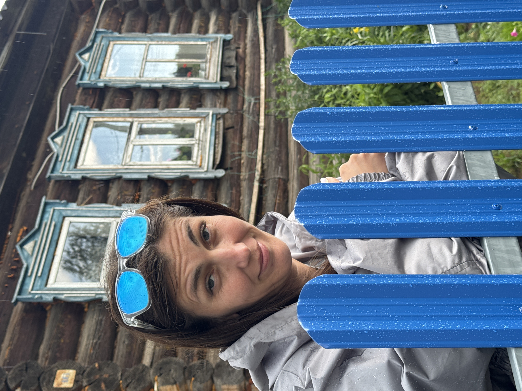In Вассята, Russia, a woman with brown hair and blue mirrored sunglasses perched on her head stands behind a bright blue corrugated metal fence. She wears a light gray, hooded, waterproof jacket, which is speckled with raindrops. The woman is looking towards the camera with a slight smile, her left hand is visible resting on the fence. Behind her, a traditional wooden log house with three windows adorned with intricate blue trim is visible. The windows reflect the sky and surrounding greenery. The overall scene suggests an outdoor setting during rainfall, possibly in a rural or suburban area.