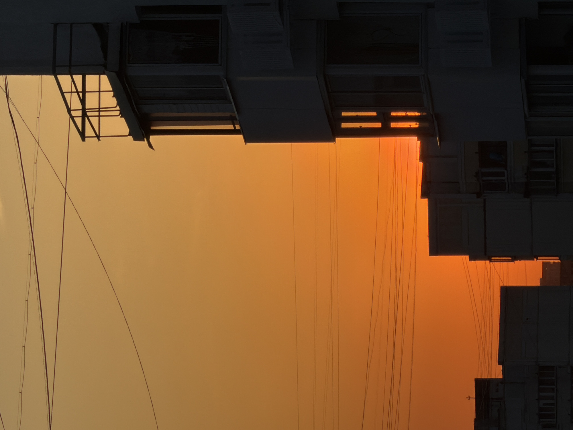An upside-down, low-angle shot depicts buildings against a hazy, orange sky, likely captured in the city of Khimki, Russia. The buildings are composed of blocky units, some with visible windows and balconies, silhouetted in dark tones. A network of power lines runs across the sky, adding linear elements to the composition. The overall atmosphere suggests either sunrise or sunset. The structures appear to be of urban residential construction. No people are visible.