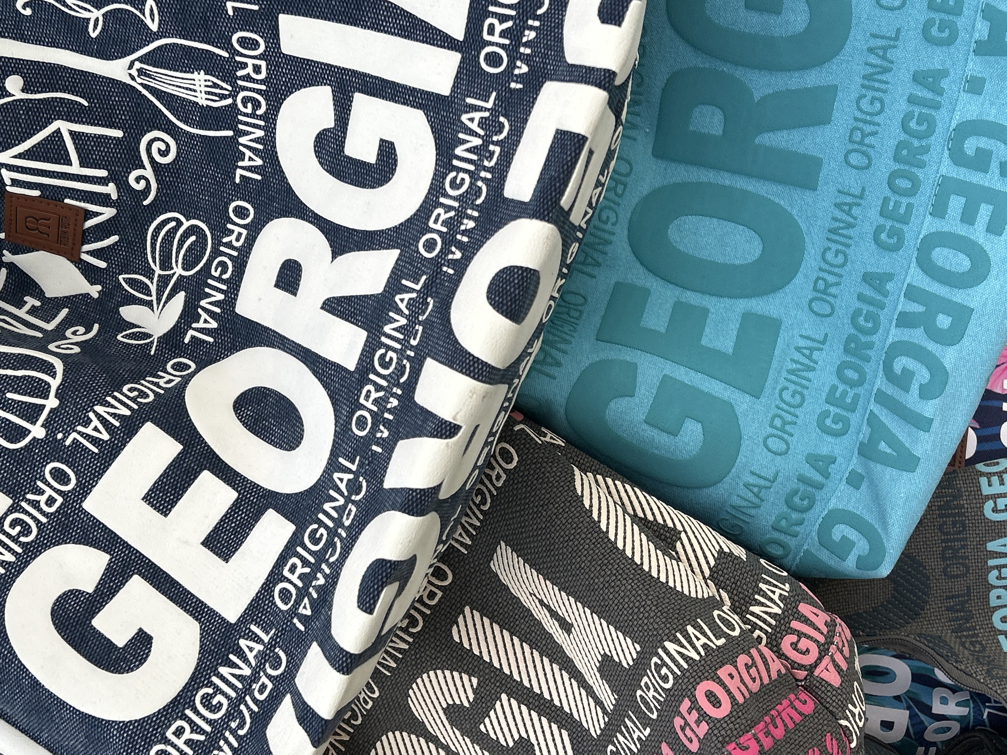 The image shows a close-up view of several fabric items, likely bags, with the word "GEORGIA" printed repeatedly in large white lettering. Some of the fabric is dark blue with white lettering, another piece is dark gray with white and pink lettering, and the bottom item is a bright teal color with raised teal lettering of "GEORGIA" and "ORIGINAL". Decorative elements like branches, swirls, and a musical instrument are also visible on the dark blue fabric. A small brown leather tag with the embossed initials "R R" is attached to the blue fabric. The items are layered on top of each other, creating a textured visual effect. There is no discernible indication of the city of Mtskheta, Georgia, in the image; it is a detail about the content of the items.