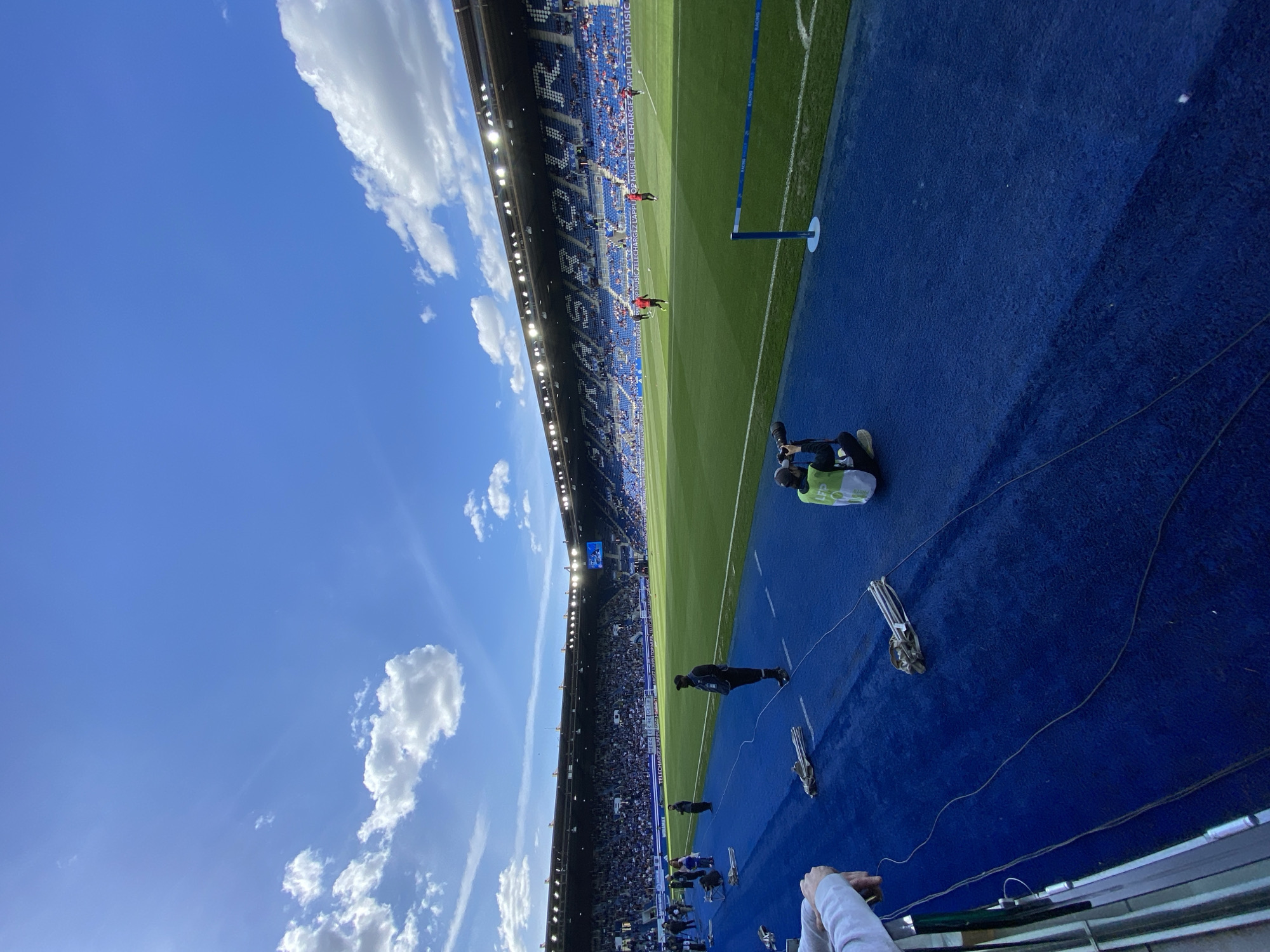 The image depicts a soccer stadium in Strasbourg, France. A portion of the blue track encircling the green soccer field is visible. Individuals are present along the track, some in official attire and others appearing to be setting up equipment. The stands are filled with spectators. The sky is blue with scattered clouds.
