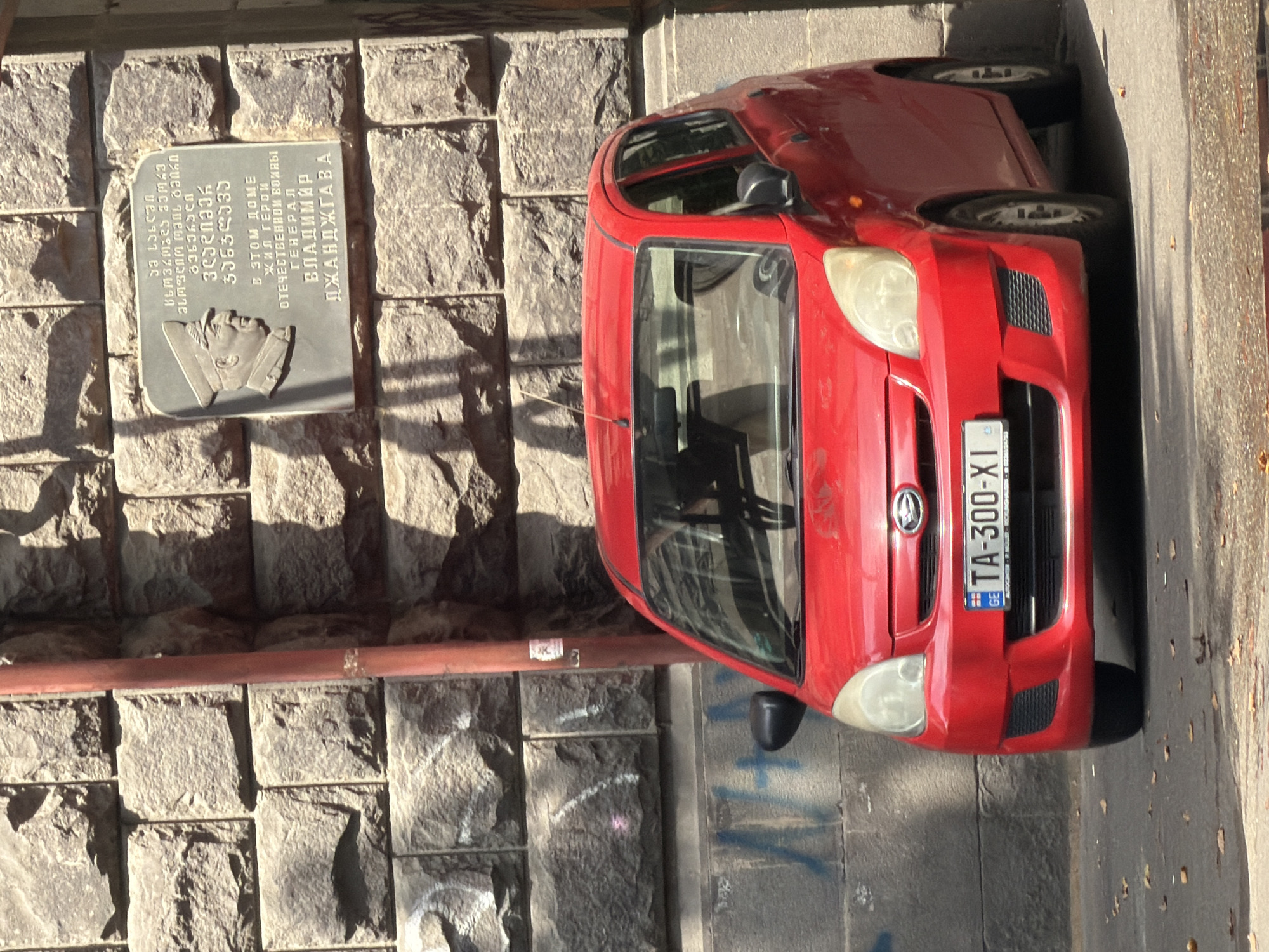In Tbilisi, Georgia, a red Daewoo Matiz is parked next to a stone wall adorned with a memorial plaque featuring a bas-relief portrait of a man in a military uniform. The plaque is inscribed with text in both Georgian and Russian, commemorating a hero of the Great Patriotic War, General Vladimir Djandjgava. The car's license plate is visible as "TA-300-XI". The sun casts shadows on the wall, indicating it is daytime. The scene appears to be on a street with fallen leaves on the ground.