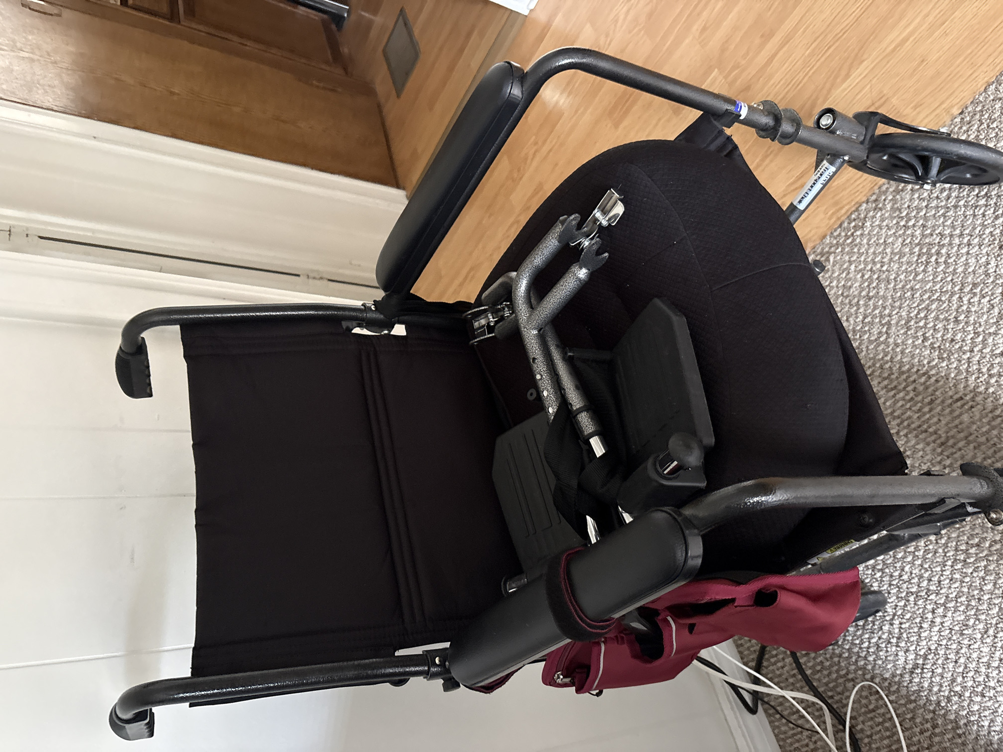 The image shows a black transport wheelchair with a maroon accessory bag attached to the back. The wheelchair is positioned on a carpeted floor next to a wooden floor. The background includes a wall with white horizontal panels and a section of wooden cabinetry. The wheelchair has black armrests, a black seat and back, and silver footplates. There is a label on the frame of the wheelchair that is partially visible. The scene appears to be indoors, in a home environment. There is no activity or event taking place, as the wheelchair is stationary. The lighting suggests it is daytime, and there are no indicators of weather or specific emotions. The location context is Bradenton, United States.