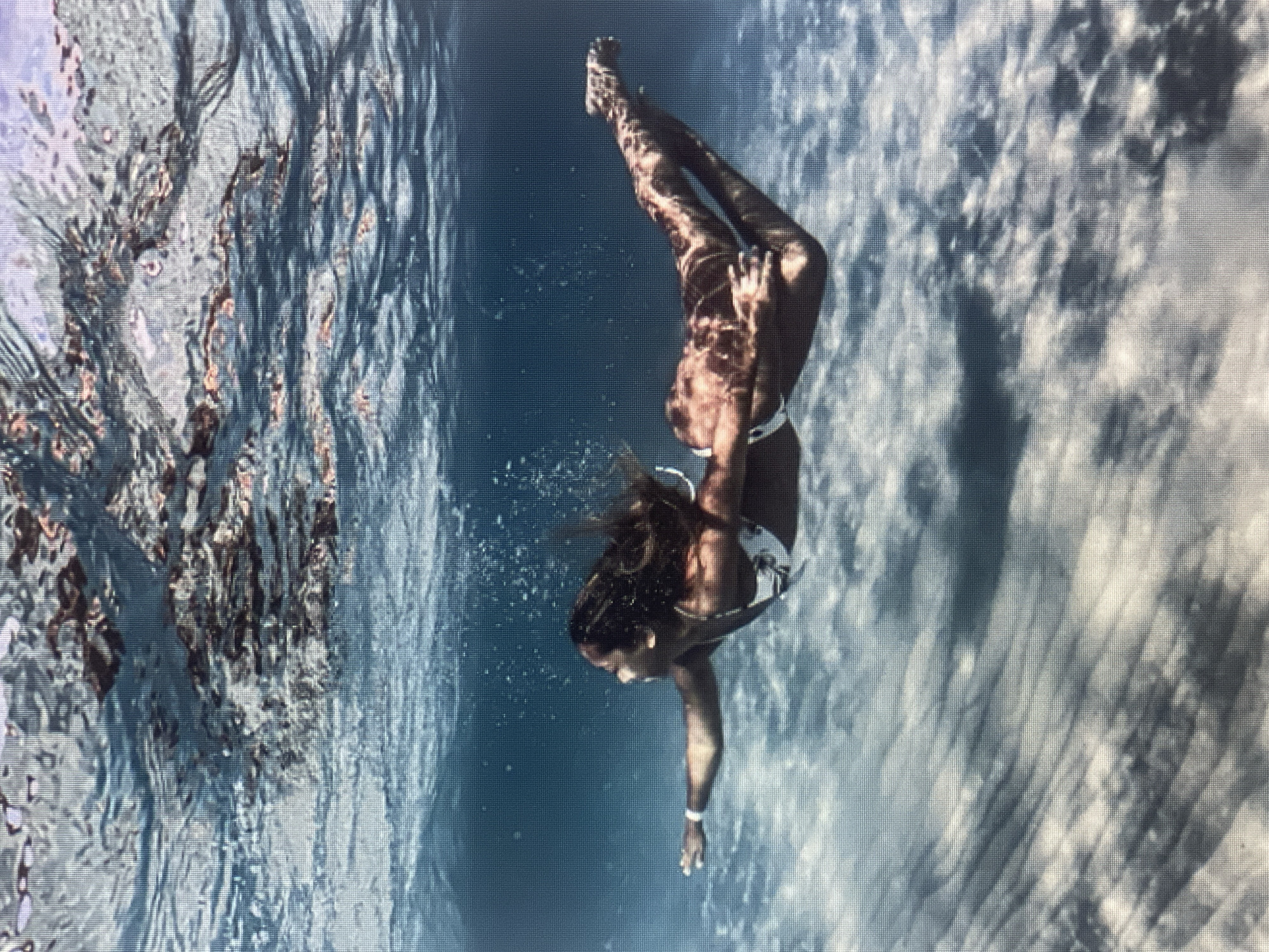 An underwater shot captures a woman swimming through clear blue water. She is wearing a white bikini and has long, dark hair that is flowing around her. The sunlight filters through the water, creating a dappled effect on her skin and the sandy bottom. The woman is positioned horizontally in the frame, with her left arm extended forward and her right arm bent at the elbow. Her body is streamlined, indicating she is actively swimming. The environment appears to be a shallow ocean or a very clear lake, with ripples of light and shadow on the seabed.