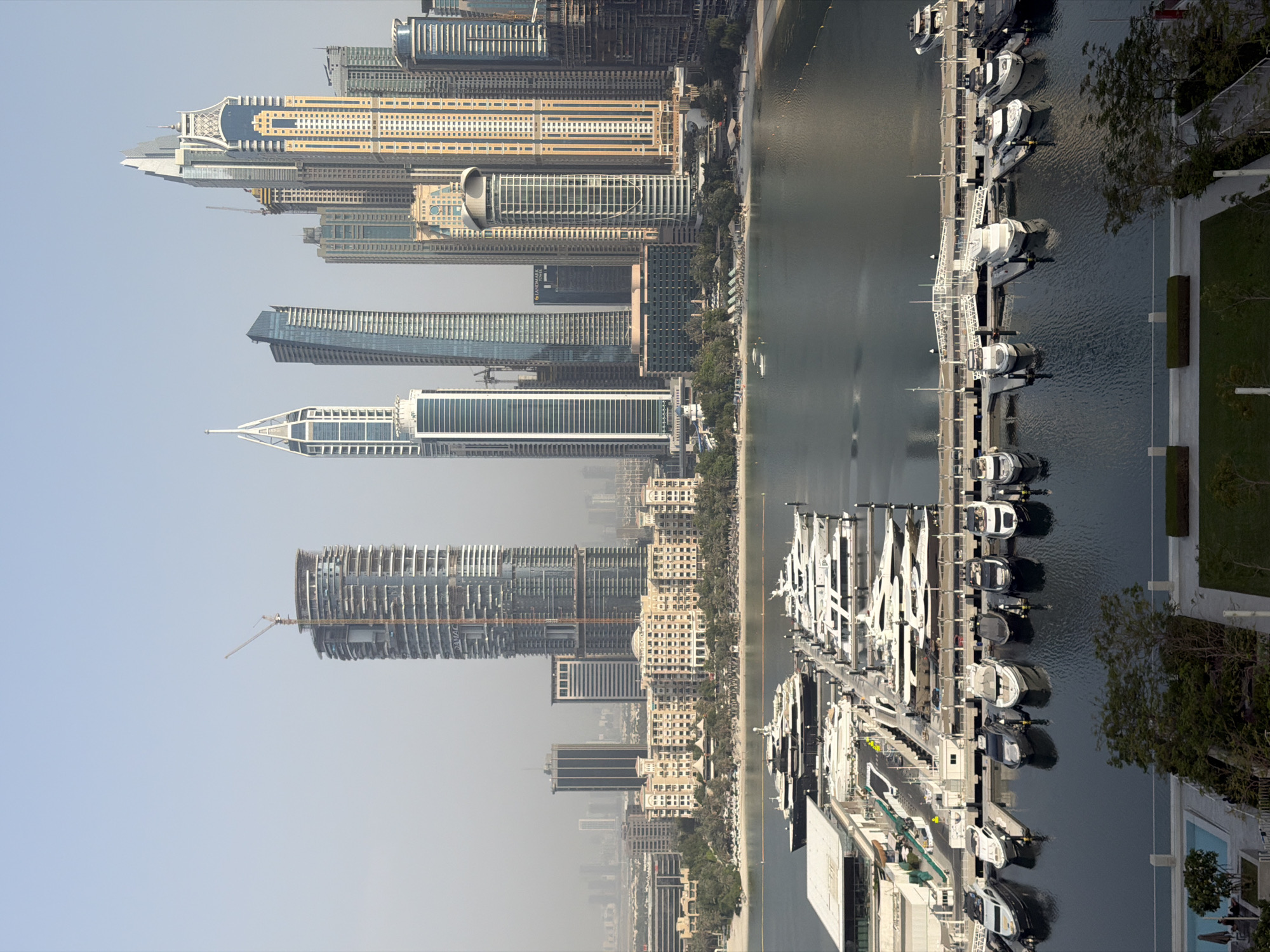 The image captures a panoramic view of the Dubai Marina, a man-made canal city in Dubai, United Arab Emirates. Dominating the skyline are numerous modern skyscrapers, characteristic of Dubai's architectural landscape. In the foreground, a marina is filled with a collection of luxury yachts and smaller boats docked along jetties. The water is calm, reflecting the surrounding buildings and sky. The background shows a beach area with palm trees and some low-rise buildings, transitioning into the vast expanse of the city's urban development. The overall scene depicts a vibrant and affluent waterfront district, showcasing a blend of residential, commercial, and leisure facilities. The sky is a pale blue, indicating clear weather conditions, possibly in the morning or late afternoon given the soft lighting.