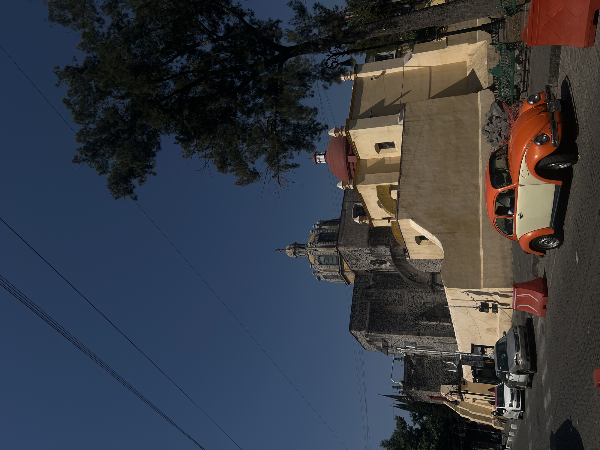 The image depicts a vibrant street scene in a historic area of Mexico City, Mexico, bathed in bright daylight under a clear blue sky. A prominent two-tone orange and cream classic Volkswagen Beetle, commonly known as a "Vocho," is parked on the right side of a cobblestone street in the foreground. Its strong shadows indicate a sunny late morning or early afternoon.
Behind the car and dominating the midground is a large, traditional church building. The lower part of the church is constructed from dark, textured stone, while its upper sections feature light, yellowish-cream plaster walls. The church showcases two distinct domes: a smaller, reddish-tiled dome on the right, and a larger, more elaborate dome with a decorative cupola or spire on the left, rising above the stone foundation.
Large, dark green trees frame the upper left portion of the image, their dense foliage contrasting with the deep blue sky. Several utility wires stretch horizontally across the sky. Further down the street, other vehicles, including a silver sedan and a white truck, are visible, suggesting an active urban environment. Green park benches and red traffic barriers are also present near the street. No direct human activity is observed, and no clear, readable text is discernible on any objects.