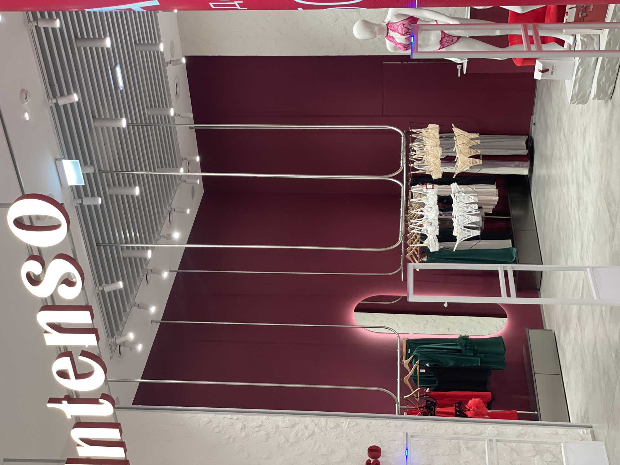 The image shows the interior of an Intimissimi store in Krasnoyarsk, Russia. The store features a burgundy wall with silver hanging rods displaying various lingerie items. The lingerie ranges in color from beige and white to dark green and red. A mannequin wearing lingerie is visible in the upper right corner of the image. The ceiling is white with rows of linear lighting fixtures. A white wall with the "Intimissimi" logo in large red letters is on the left. The flooring appears to be light gray or white. A partial view of a shelving unit is visible in the bottom right corner. The overall ambiance is modern and clean.