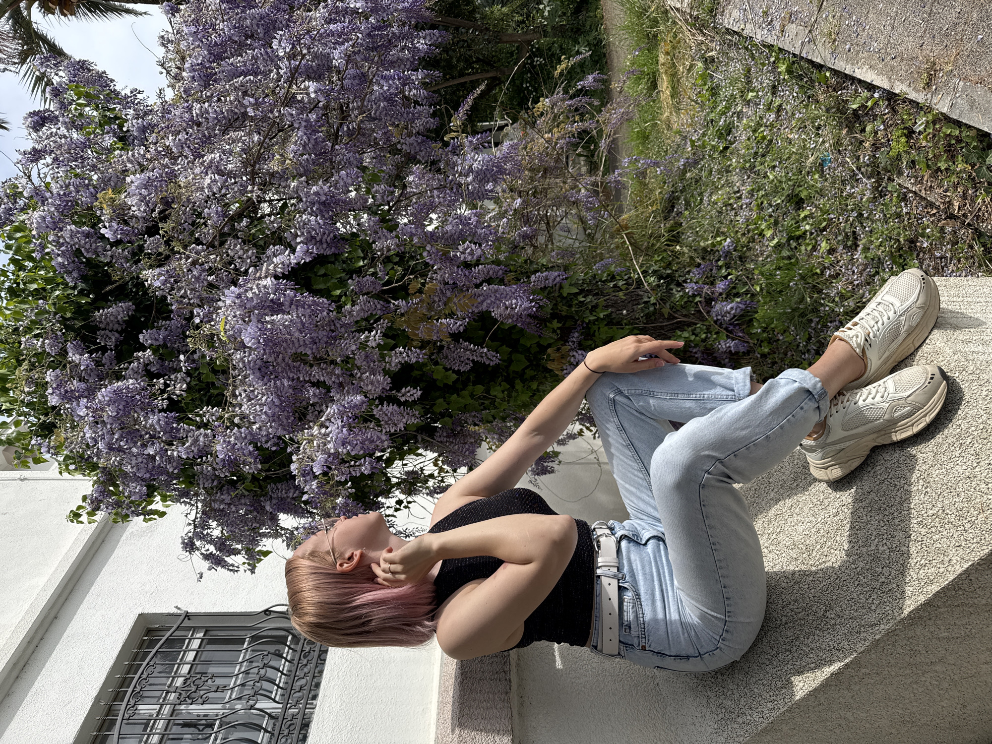 A young woman with blonde and pink-tinted hair is seated on a concrete ledge in Bar, Montenegro. She is wearing a black tank top, light blue jeans with a white belt, and white sneakers. The ledge appears to be part of a building's exterior, near a window with decorative bars. Behind her, a wisteria tree blooms with an abundance of purple flowers, and there is also green vegetation visible. The scene is bathed in sunlight, indicating it is daytime. The environment suggests an urban setting with natural elements integrated into the architecture or landscaping.