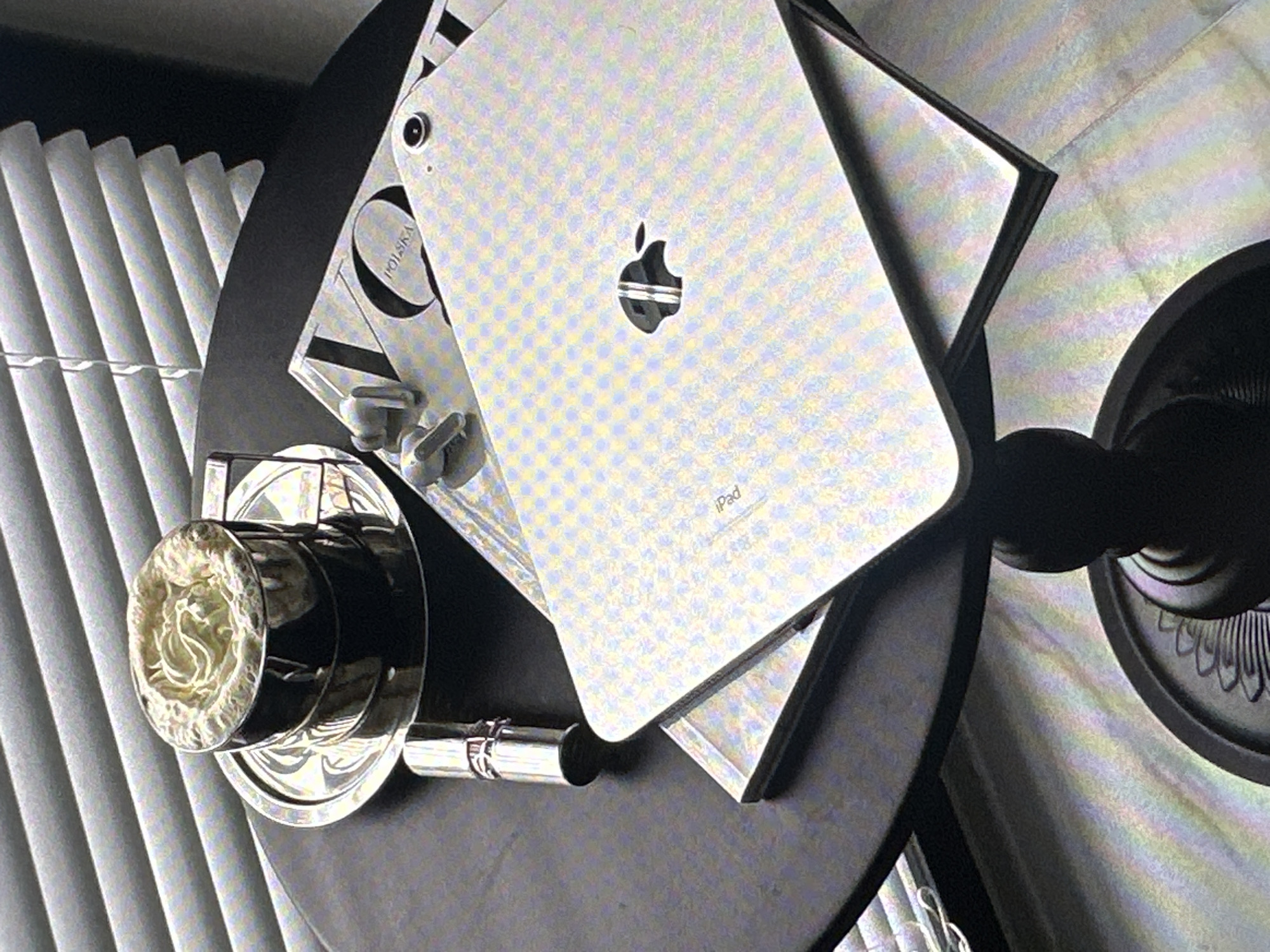 The image shows an iPad on a black round table, along with a magazine, a perfume bottle and an ornate cup with a decorative lid. The table is near a window with white blinds. The location is not identifiable as Yerevan, Armenia based on visual information alone.