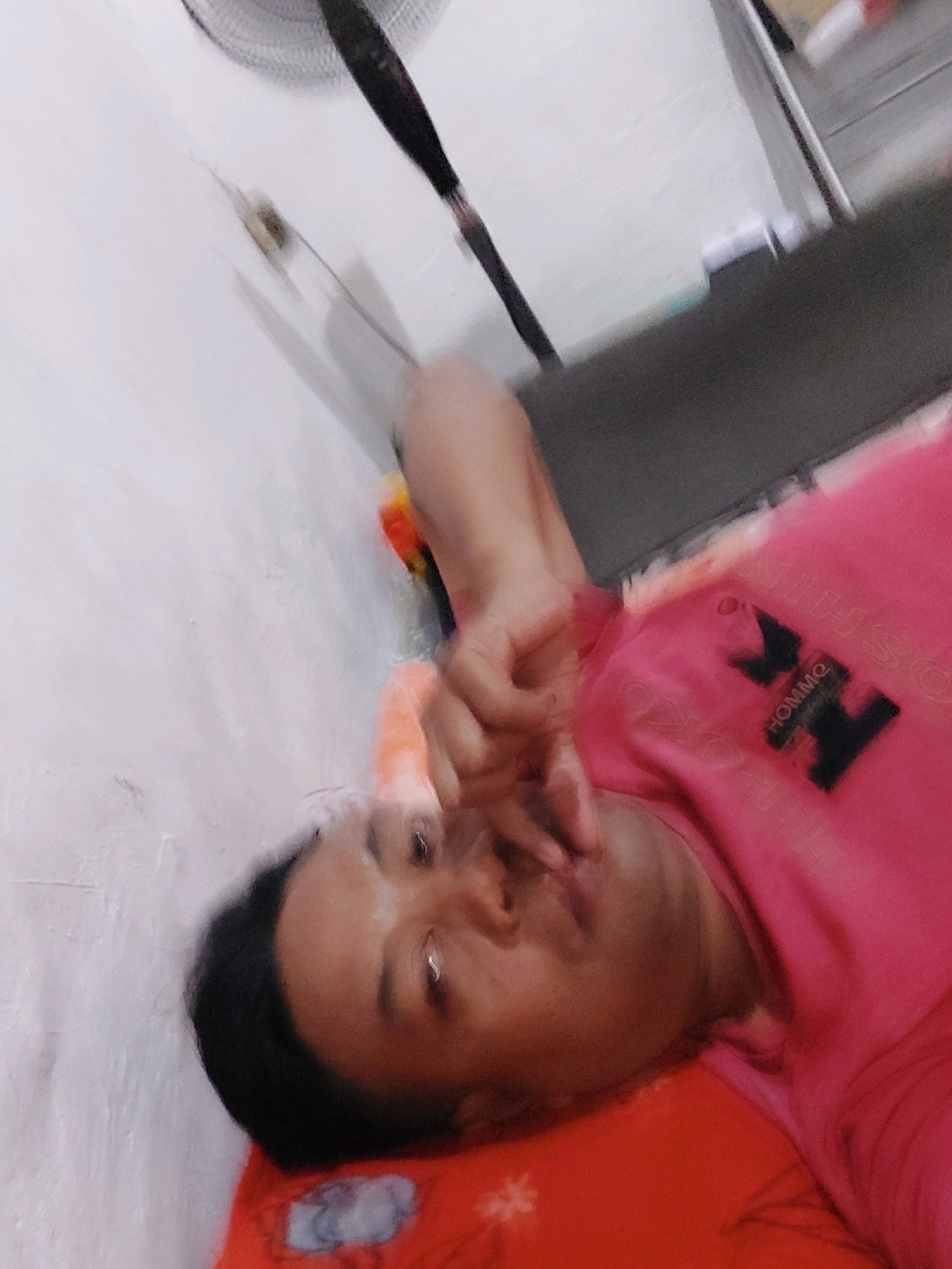 The image shows a person lying down on a red surface, possibly a bed, in a room. The person is wearing a pink shirt with a black logo that appears to say "HOMME". Their face is partially obscured by their hand, and they are looking upwards. The background is a white wall with a dark object, possibly a fan, mounted on it. The lighting in the room is dim. The scene appears to be an indoor setting, and there are no specific visual cues that can definitively place the location in Sarangan, Indonesia. The image is slightly blurry, suggesting it may have been taken with a mobile phone camera.