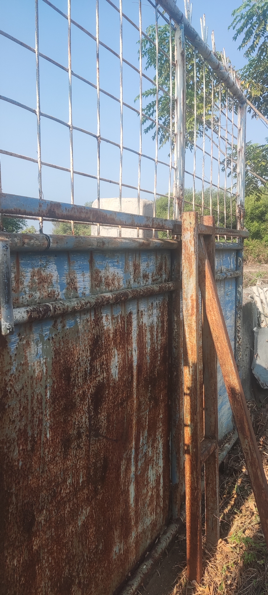 An outdoor, medium-shot, vertical photograph captures a rusted metal gate against a bright blue sky. The gate, made of vertical bars on the top section and a solid panel on the bottom, exhibits significant rust and peeling blue paint. A vertical, rusty metal post stands to the right of the gate, with a diagonal rusty support beam attached to it. Green foliage and trees are visible behind the gate and to the right, suggesting an outdoor environment, possibly rural or overgrown. The lighting indicates it is daytime, with sunlight casting shadows on the metal structures. The location is specified as Gebang, Indonesia.