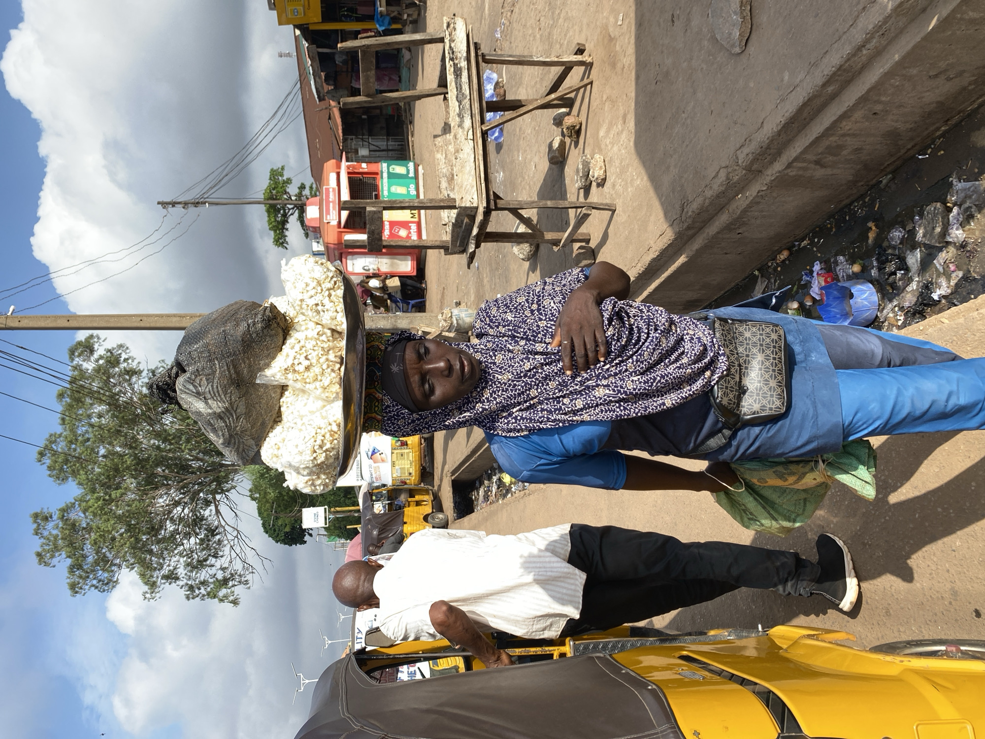 In Sabon Tasha, Nigeria, a woman walks along a street carrying a large tray of white, puffed food items, possibly popcorn, balanced on her head. She is wearing a blue tunic, bright blue trousers, and a patterned headscarf. A small, patterned cross-body bag is secured at her waist. To her left, a man in a white shirt and black trousers walks away from the camera, his hand on the back of a yellow auto-rickshaw. The street is lined with a concrete drainage ditch filled with trash and debris. In the background, there are market stalls, including a red booth with signs for "Airtel" and "Glo," and a wooden stall with a makeshift table. Power lines crisscross the sky, and a large tree with full foliage stands prominently. The sky is partly cloudy.