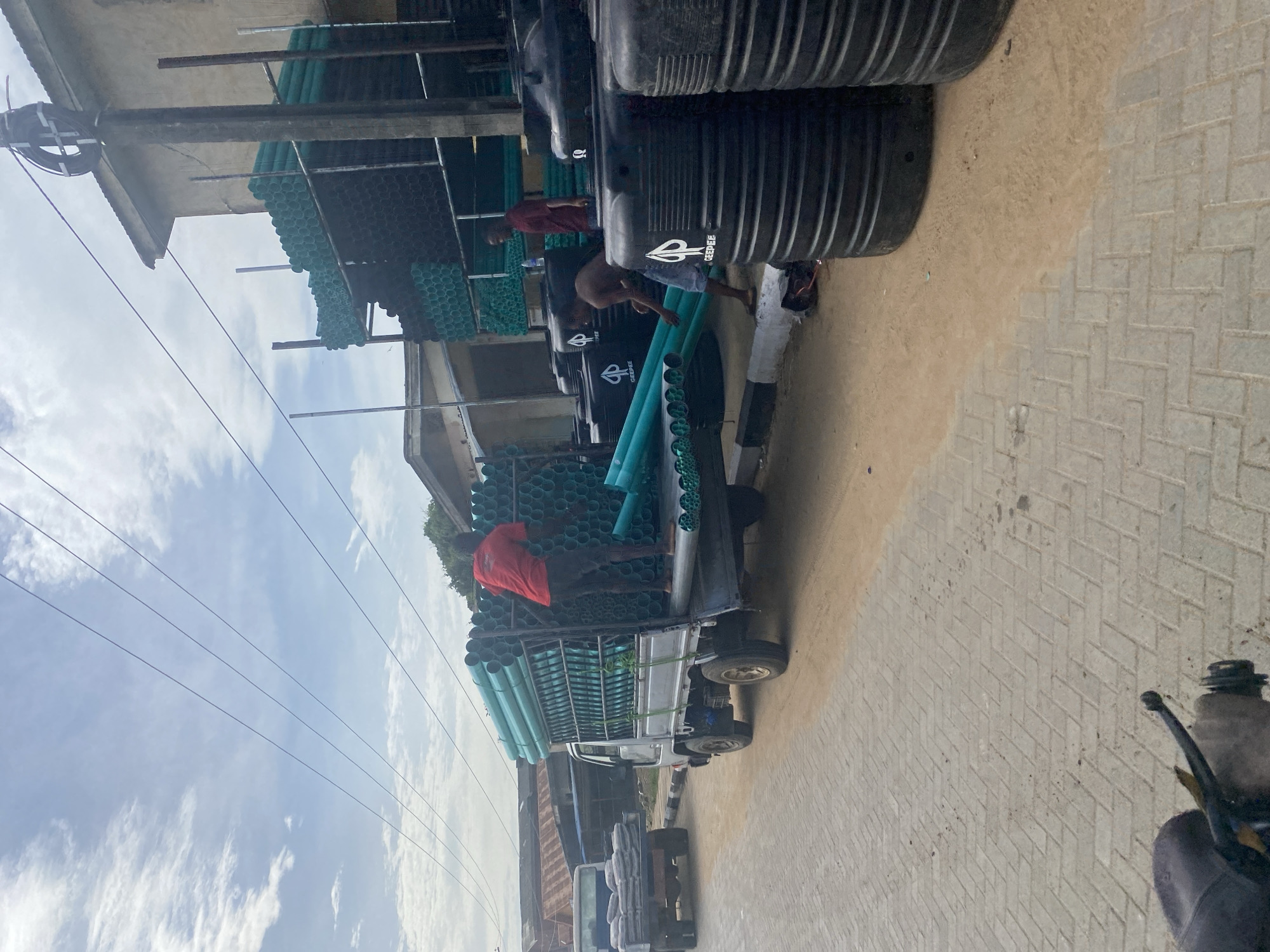 The scene depicts a street in what appears to be a residential area. A truck is loaded with green pipes and several black water tanks. Two individuals are visible near the truck, seemingly involved in loading or unloading. The surrounding environment includes buildings, power lines, and a brick-paved road. The city and region are not identifiable from the provided details.