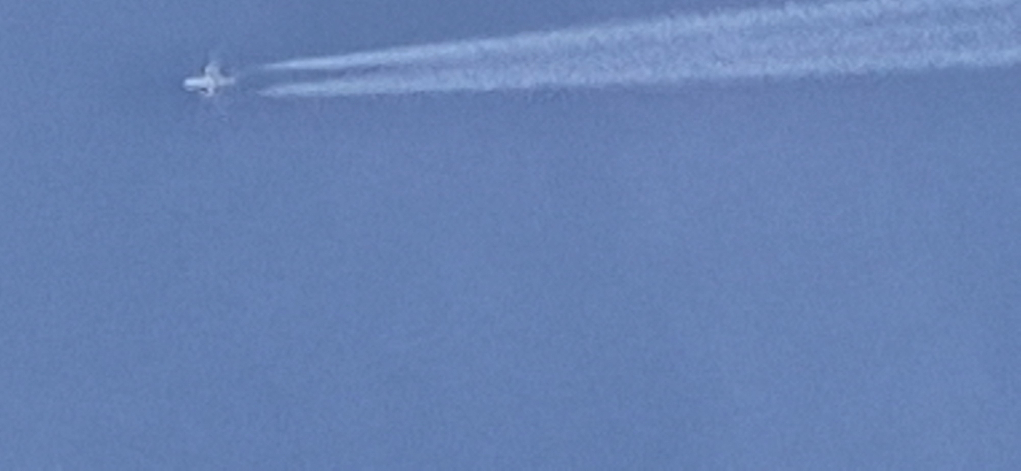 An airplane, leaving a contrail, is visible in the upper right quadrant of a clear blue sky, likely over the city of Mainz, Germany. The airplane appears to be a small, white jet with its wings extended. The contrail is a thick, white trail that extends downwards and to the left from the airplane, gradually dissipating into the sky. The rest of the image is dominated by the uniform blue of the sky, with no other discernible objects or features. The image quality is somewhat blurry, suggesting it may have been taken from a distance or with a low-resolution camera.