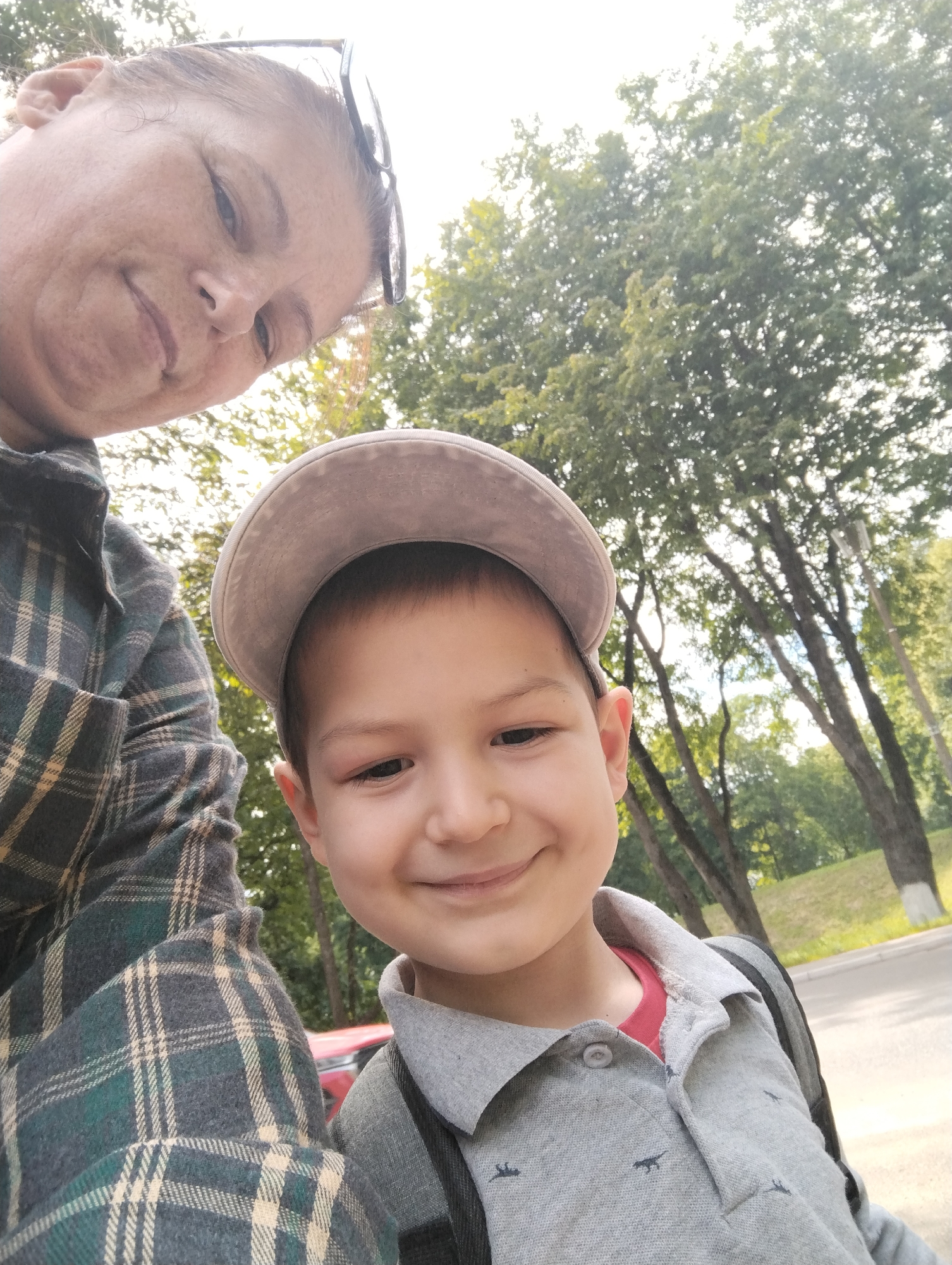 A selfie is taken outdoors, possibly in a park or along a tree-lined street. In the foreground, two individuals are visible. On the left, a woman's face and shoulder are shown from a downward angle. She has glasses resting on her head and is wearing a dark green and cream plaid shirt. To the right of the woman, a young boy is prominently featured, smiling at the camera. He is wearing a light grey polo shirt with small dark animal prints and a grey baseball cap. The background consists of lush green trees, suggesting a natural or park-like setting. The lighting indicates it is daytime. The scene does not contain any specific identifiable landmarks of Kingisepp, Russia.