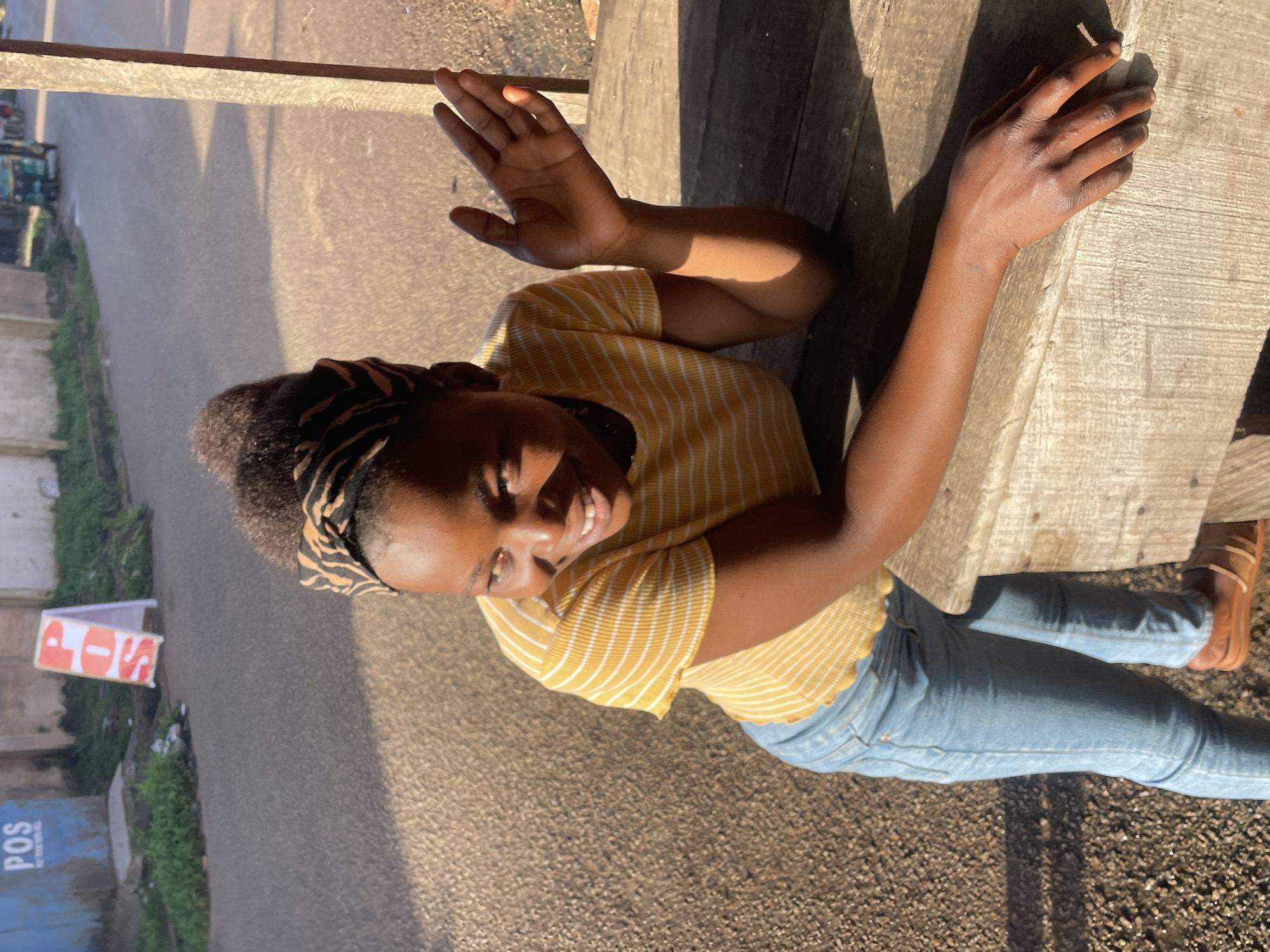 A young Black female with dark skin and curly hair is leaning on a wooden table. She is wearing a yellow and white striped t-shirt, light blue jeans, and brown sandals. A black and yellow patterned headwrap is on her head. Her left arm is resting on the table, and her right arm is raised with her palm facing forward. She is looking to her left and smiling. Sunlight is casting shadows across the scene. In the background, a road is visible with some greenery and buildings. A blue POS sign is on the left side of the frame, and a white A-frame sign with orange lettering is also visible on the road. A trishaw can be seen in the distance on the road. The overall setting appears to be an outdoor, urban environment.