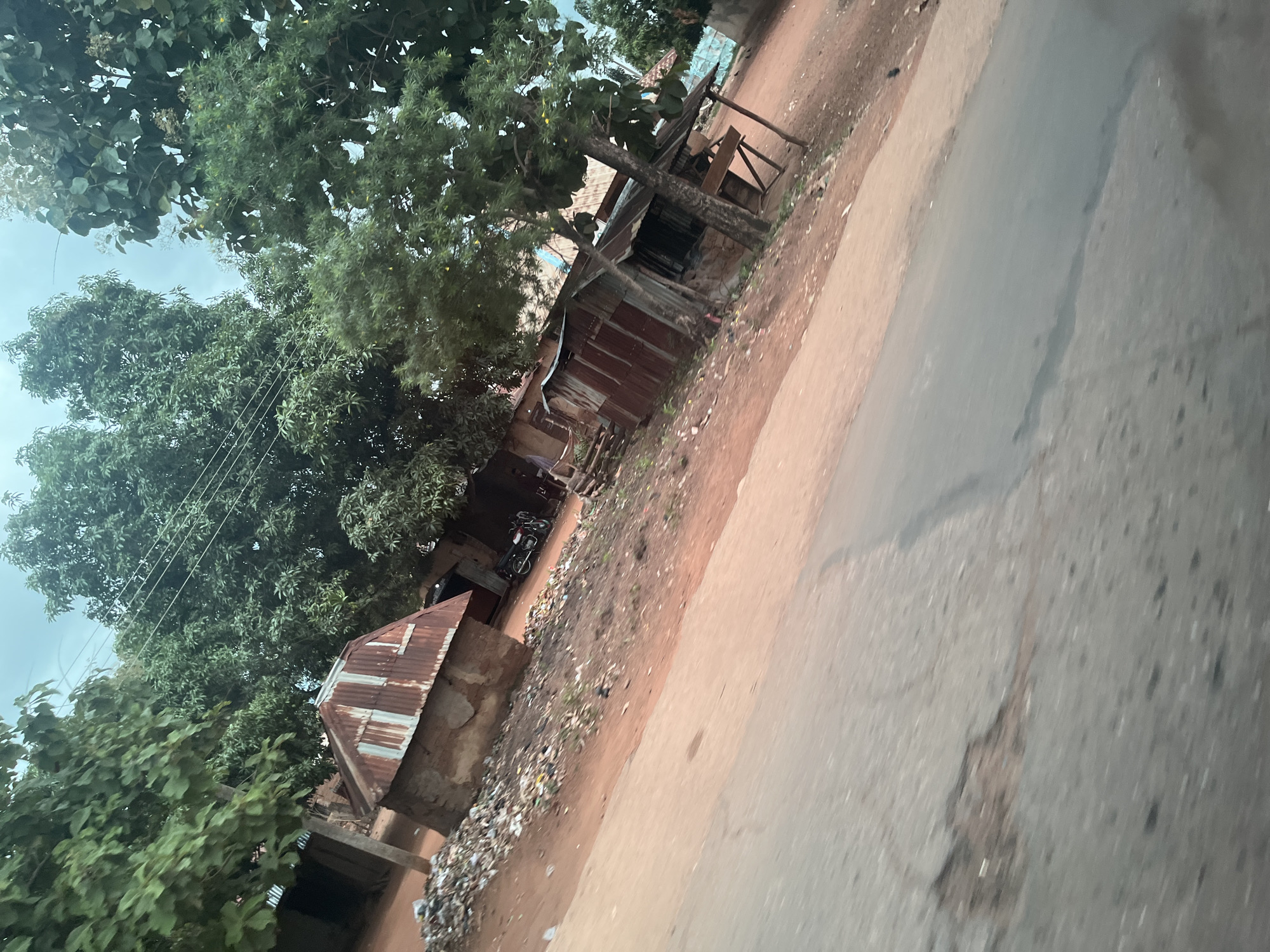 The image shows a street scene in Karshi, Nigeria. The foreground is dominated by a cracked asphalt road, with the right side of the road exhibiting a reddish-brown dirt surface. Along the left side of the road, there is a pile of debris and trash. Behind the trash pile, a small, circular hut with a rusted, corrugated metal roof is visible. Further back, several structures with corrugated metal roofs, appearing to be shacks or small buildings, line the dirt road. There are trees with lush green foliage scattered throughout the scene, some with electrical wires running through them. A motorcycle is parked near one of the structures. The sky is overcast.