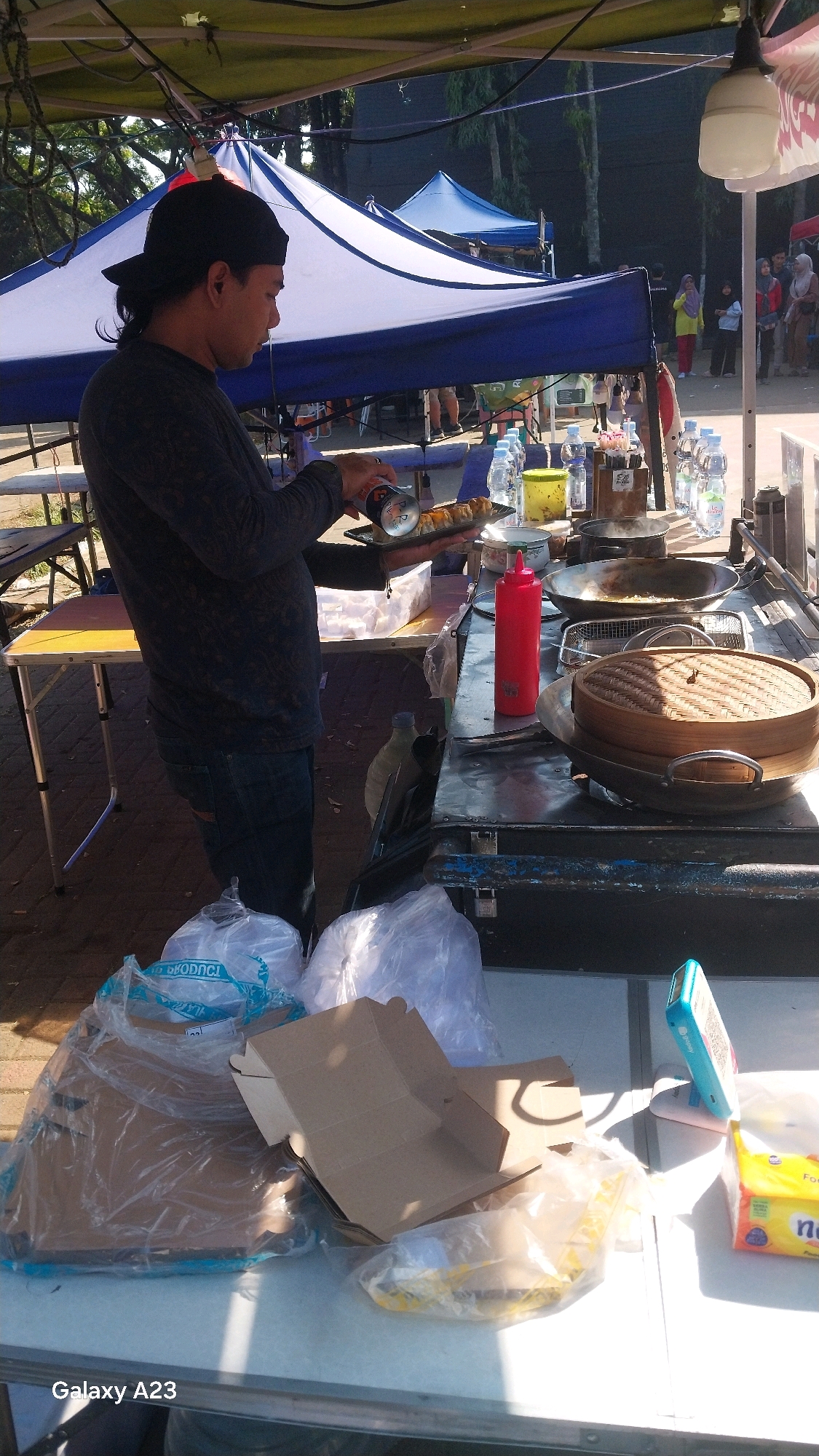 This is a medium shot, eye-level, outdoor image taken on a sunny day. The foreground is dominated by a white folding table laden with various items including plastic bags filled with unknown contents, cardboard food packaging, and a yellow package of what appears to be tissues or napkins. To the left, the "Galaxy A23" watermark is visible. The midground features a food stall with a man wearing a dark long-sleeved shirt, dark pants, and a black baseball cap, holding a can over a plate of food. The stall is equipped with cooking utensils including a wok, a steamer basket, and bottles of water. In the background, several tents are visible, one blue and white, and another smaller blue tent. People are seen milling around in the background. The overall setting suggests a street market or outdoor food vendor area. Based on the presence of the "Galaxy A23" watermark, it is probable that the image was captured in Malang, Indonesia, as this is a common feature on images taken by Samsung devices, and the environment is consistent with typical street scenes in Indonesian cities.