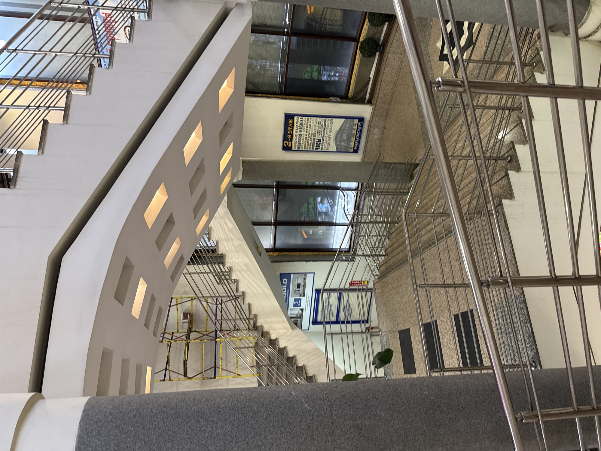 The image depicts an indoor staircase scene, likely in a commercial or public building, possibly in Moscow, Russia. The stairs have metal railings and white steps, and the architecture features a curved wall with recessed lighting. Signage in Russian (with the word "GOLD" and "MAR'INSKIY") indicates the building's function, likely containing businesses selling gold jewelry, women's and men's clothing, watches, and souvenirs. The presence of scaffolding suggests ongoing maintenance or renovation work. The overall environment appears modern and well-lit.