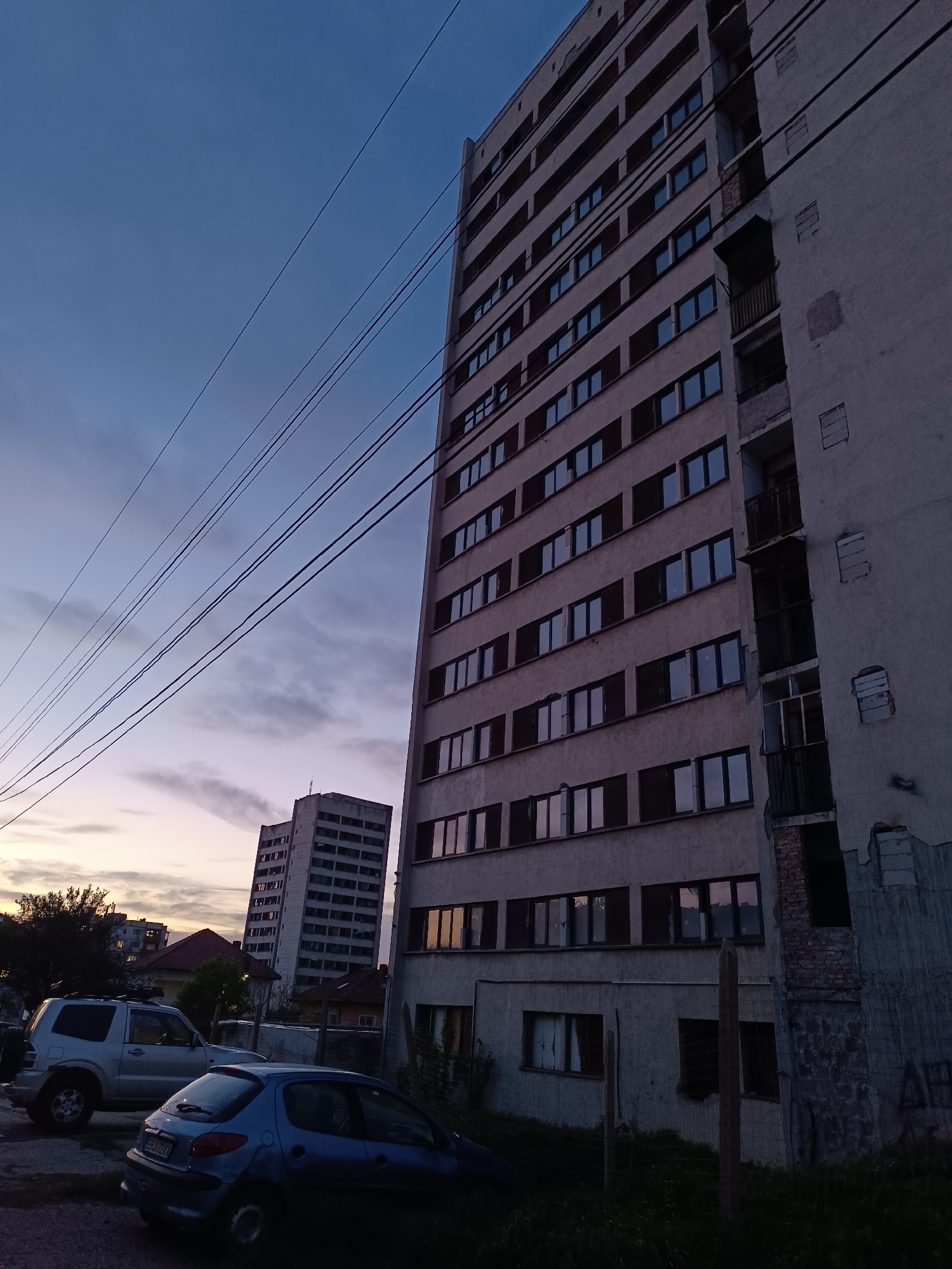 The image depicts an urban or semi-urban scene at dusk or dawn, characterized by a twilight sky transitioning from deep blue overhead to lighter purples and pinks near the horizon. Electrical power lines stretch across the sky.
The primary subject is a tall, multi-story concrete apartment building occupying the right side of the frame. Its facade features numerous rectangular windows, some reflecting the dim sky. The building shows significant signs of wear and tear, with sections of its right side appearing damaged, revealing exposed brickwork and concrete. In the middle distance on the left, another similar, though slightly smaller, concrete building stands against the colorful horizon.
In the foreground and midground, two cars are parked. On the left, a silver SUV, possibly a Mitsubishi Pajero, is partially visible. Closer to the center, a blue Peugeot 206 hatchback is parked. Its rear license plate, "CB 3172 KX," is clearly legible, indicating a Bulgarian registration (Sofia City). Various trees, bushes, and overgrown grass are scattered around the vehicles and at the base of the buildings. Faint, dark graffiti or markings are barely visible on the lower right wall of the main building, though largely illegible. The scene is devoid of people or overt activity, conveying a quiet, contemplative atmosphere in the fading or emerging light.