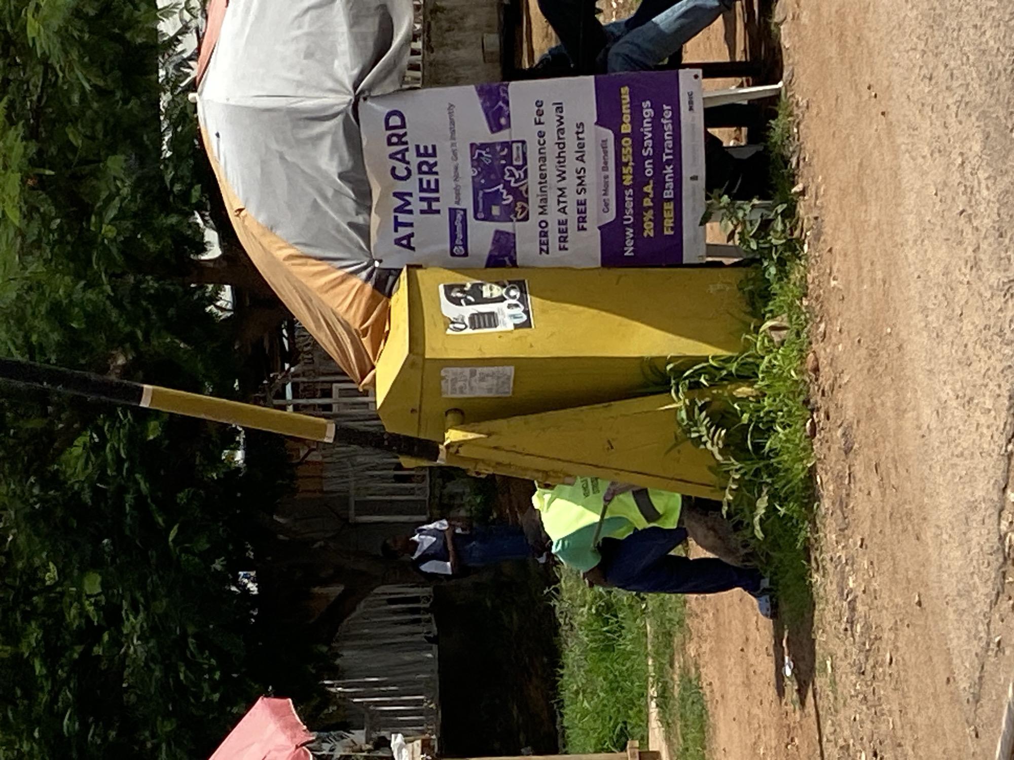 This is a medium shot of an outdoor scene in Nigeria, likely Gura Top. The image is oriented vertically. In the foreground, a yellow kiosk-like structure stands on a dirt path surrounded by greenery. A person wearing a bright green reflective vest and dark pants is crouched down next to the kiosk, seemingly working. Their face is not clearly visible. Behind them, a large white and tan canopy is set up, partially obscuring a brick wall and some trees.
Attached to the kiosk is a purple and white banner advertising "ATM CARD HERE" with the Palmpay logo. The banner lists benefits such as "ZERO Maintenance Fee," "FREE ATM Withdrawal," and "FREE SMS Alerts." It also promotes a "New Users N5,550 Bonus," "20% P.A. on Savings," and "FREE Bank Transfer." There are images of bank cards on the banner.
A black and yellow striped pole, possibly part of a barrier or gate, extends across the left side of the image. The scene appears to be outdoors during the daytime, with bright sunlight casting shadows, suggesting fair weather. The overall atmosphere is one of a busy, functional outdoor space, likely a point of service.