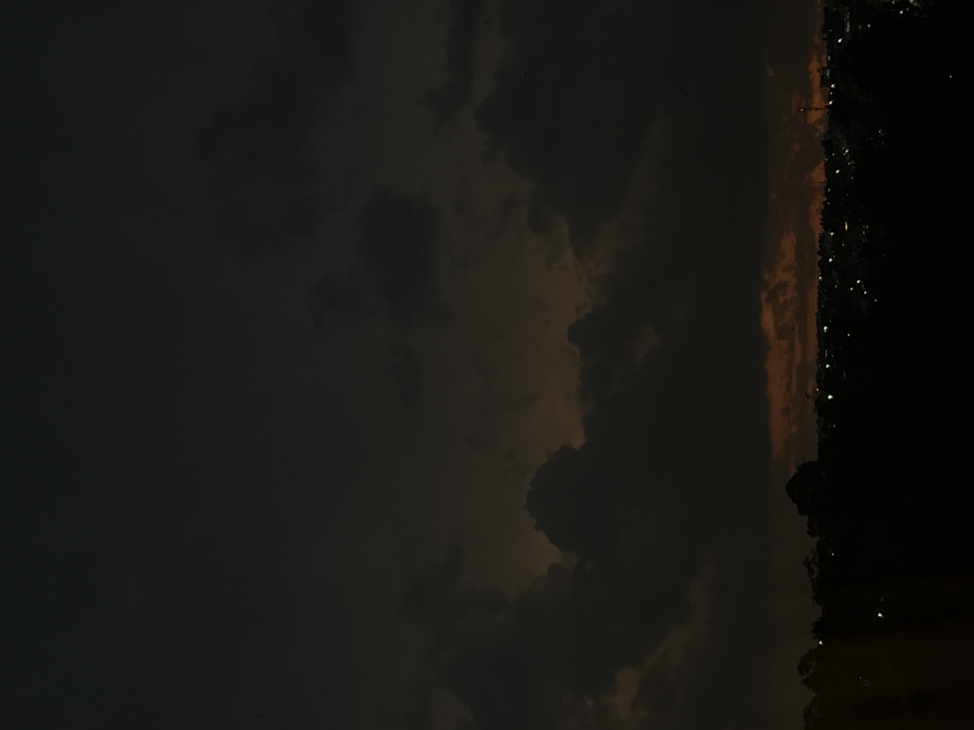 The image shows an evening or night scene, with a dark sky filled with clouds. On the right side of the image, there is a faint skyline with scattered lights, possibly indicating a city. Given the prompt's mention of Mexico City, Mexico, it's plausible this is an outlying area of the city or a view from a high vantage point overlooking the cityscape. However, the darkness obscures precise details about the environment and any specific activities or subjects. The primary focus is on the cloudy sky and the distant urban lights.