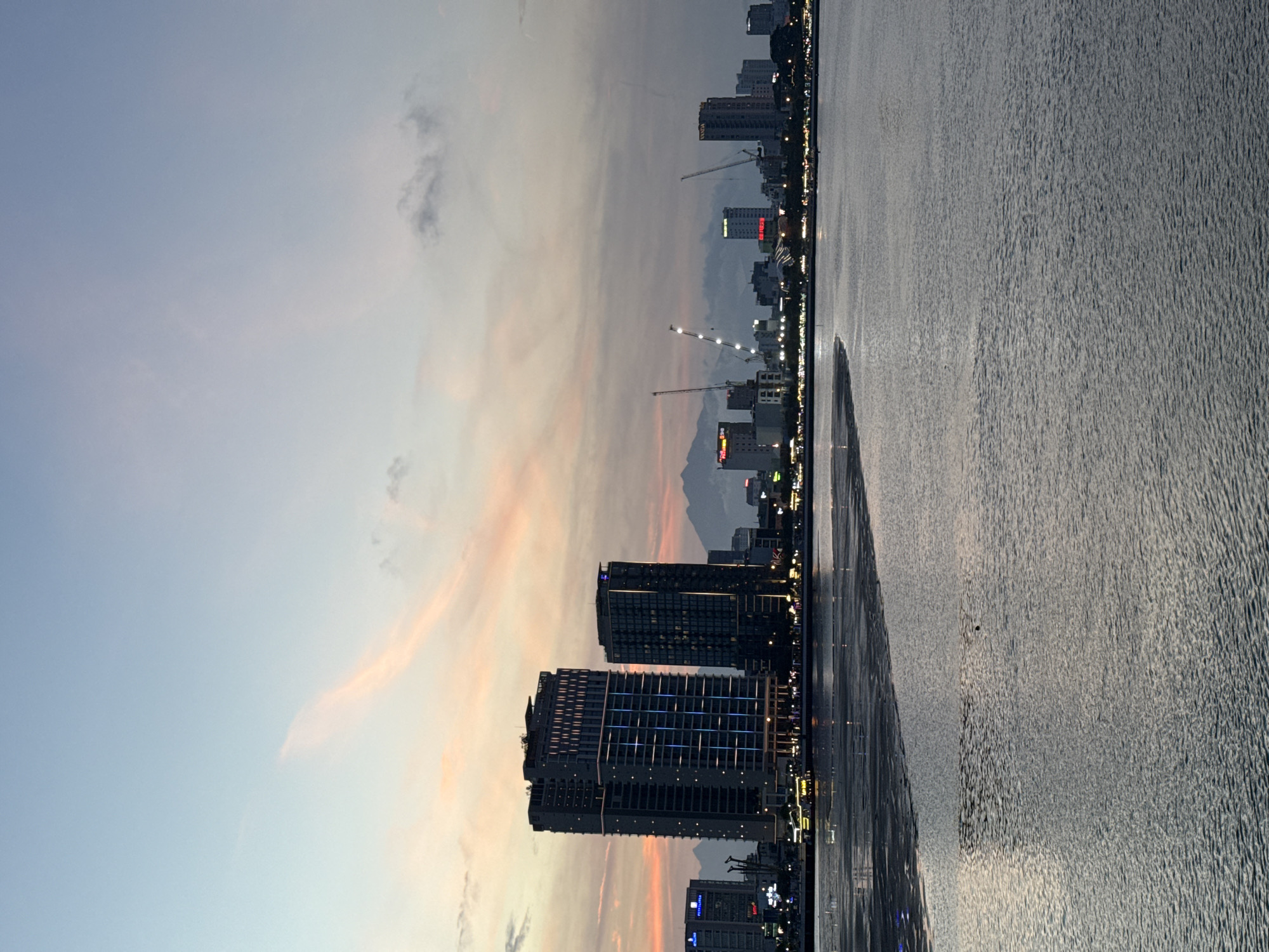 The image captures a scenic view of the city of Da Nang, Vietnam, at dusk, likely from across the Han River. The sky displays a gradient of soft blues, pinks, and oranges, indicative of a sunset. Dominating the skyline are several modern high-rise buildings, two of which are particularly prominent and illuminated with vertical lines of blue and yellow lights. In the middle ground, the city's shoreline is visible, with a stretch of sandy beach or tidal flats leading to the water. Several boats are present on the water, some appearing to be illuminated. Construction cranes are also visible among the buildings, suggesting ongoing development. In the distance, a range of mountains forms a hazy backdrop against the twilight sky. The foreground is occupied by the calm waters of the river, reflecting the ambient light and the distant city lights. The overall atmosphere is serene and captures the transition from day to night in a coastal urban environment.
