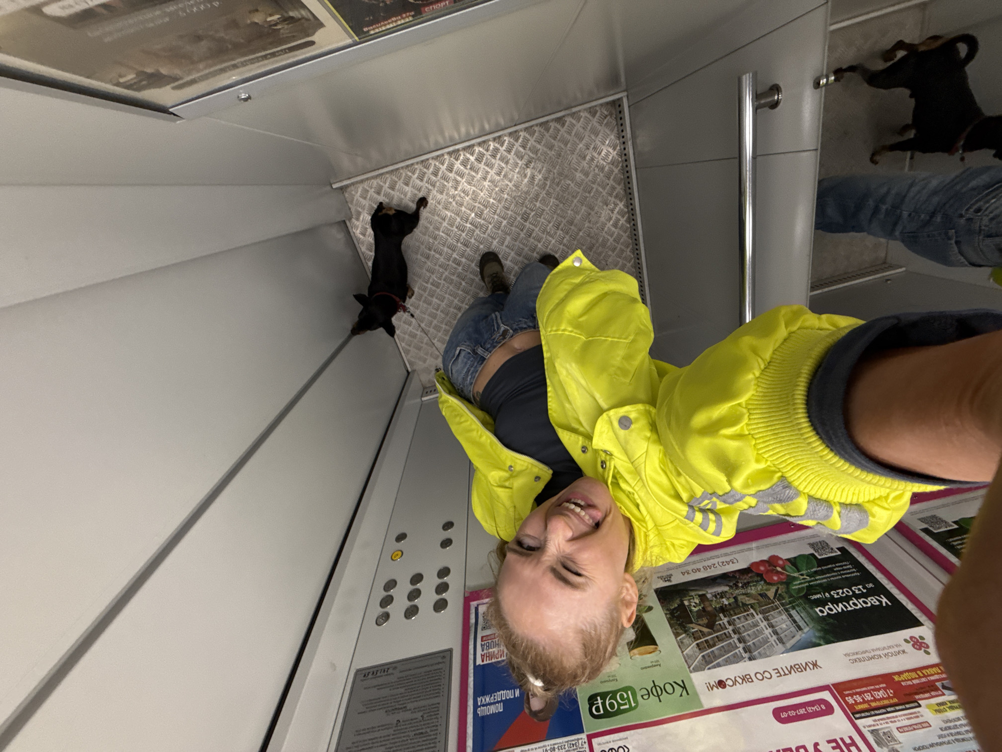 Here is a concise description of the image:
The image appears to be a selfie taken by a woman in an elevator. She is wearing a bright yellow jacket and blue jeans. A small black dog with a red collar is lying on the elevator floor near her feet. The elevator has advertisements posted on the walls in Russian, including promotions for apartments, coffee, and a holiday sale. There are also buttons on the elevator's control panel. Based on the advertisements, this image was likely taken in Perm, Russia.