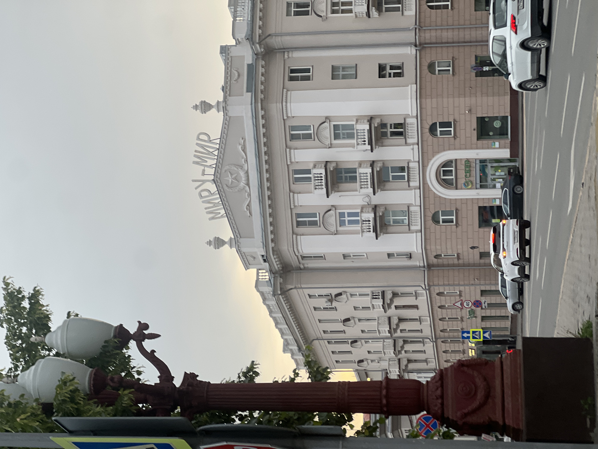 The image depicts a multi-story building in Kazan, Russia. On top of the building, the Cyrillic letters "МИРУ-МИР" are visible, translating to "Peace to the World." The building's architecture includes balconies and decorative elements, and it has a branch of Sberbank on the ground floor. A street with passing cars is visible to the right of the building, along with street signs and a sidewalk. In the foreground, there's a lamppost with globe-shaped lamps, partially obscured by foliage. The sky is overcast.