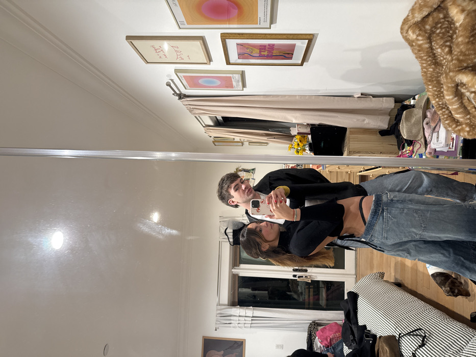 A mirrored image of a couple is visible. The woman, with long brown hair, wears a black top and blue jeans. A man, wearing a black jacket and yellow shirt, stands beside her. They are taking a selfie with a phone. The room contains framed artwork on the wall, a window with curtains, a striped sofa, and various personal items.