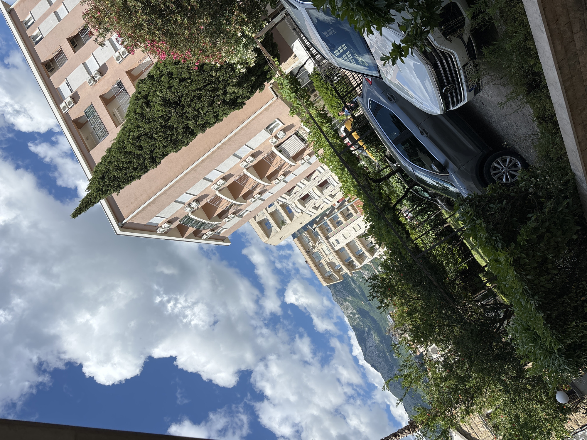 This image depicts an outdoor scene in Budva, Montenegro, with a focus on residential buildings, vehicles, and natural elements under a partly cloudy sky.
In the foreground, two cars are parked on a sloped surface. The car on the right is a white Volkswagen, identifiable by its grille and emblem, with a license plate visible. The car to its left is a gray SUV. Lush green foliage, including bushes and trees, frames the lower part of the image, suggesting a garden or landscaped area.
Behind the cars, a black metal fence runs horizontally, partially obscured by vegetation. A trellis structure with green vines is also present, adding to the garden-like atmosphere. A person, appearing to be a child, can be seen briefly near the fence, wearing a blue shirt.
Dominating the mid-ground are several multi-story residential buildings. The most prominent is a tall, light pink building with multiple balconies, each adorned with railings. Air conditioning units are visible mounted on the building's facade. To the left and behind this building, other apartment complexes with varying architectural styles are visible, featuring balconies and windows. A tall, slender, dark green conifer tree stands prominently next to the pink building, reaching towards the sky. Pink and white flowering bushes add touches of color to the right side of the scene.
In the background, a majestic mountain range rises under a bright blue sky dotted with fluffy white clouds. The slopes of the mountains are covered in vegetation, and some rocky outcrops are visible. The overall impression is a sunny day in a Mediterranean coastal city, with a blend of urban development and natural beauty.