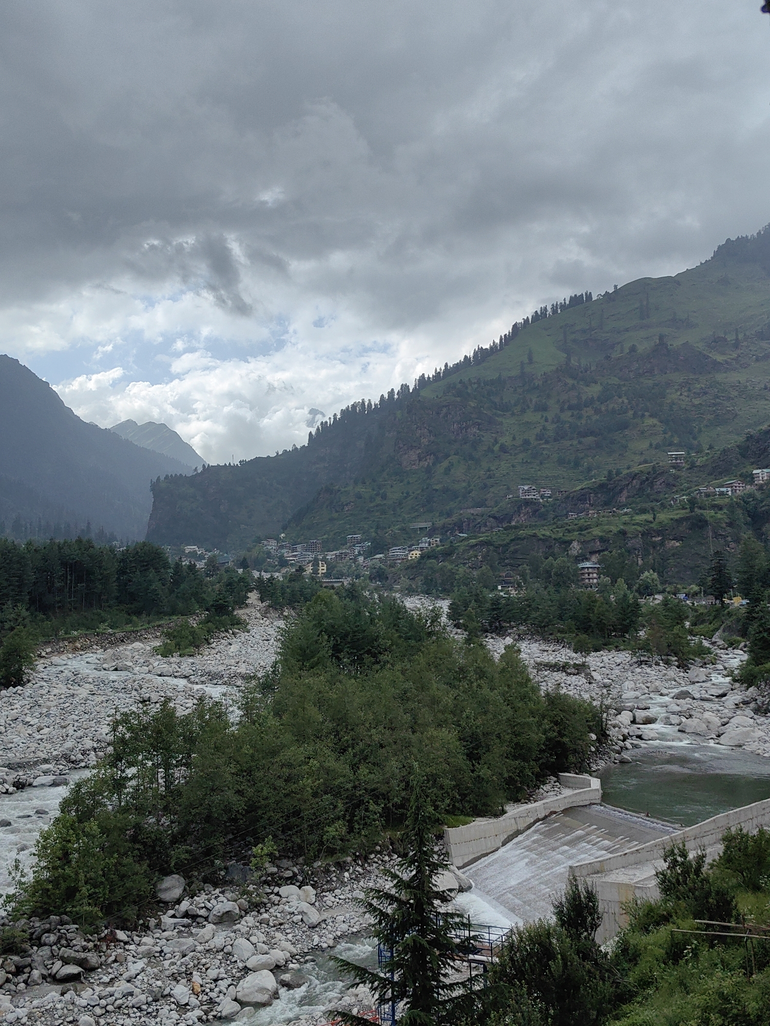 A view of a mountainous landscape with a river flowing through it, located in or near the city of Kullu, India. The scene is dominated by steep, tree-covered hillsides and a wide riverbed filled with smooth, gray stones. A concrete dam structure with water flowing over it is visible in the lower right portion of the image. In the distance, several buildings are nestled into the hillsides, indicating a settlement. The sky is overcast with heavy gray clouds, suggesting a cool or rainy day. The overall impression is one of a natural, somewhat rugged environment with evidence of human habitation.