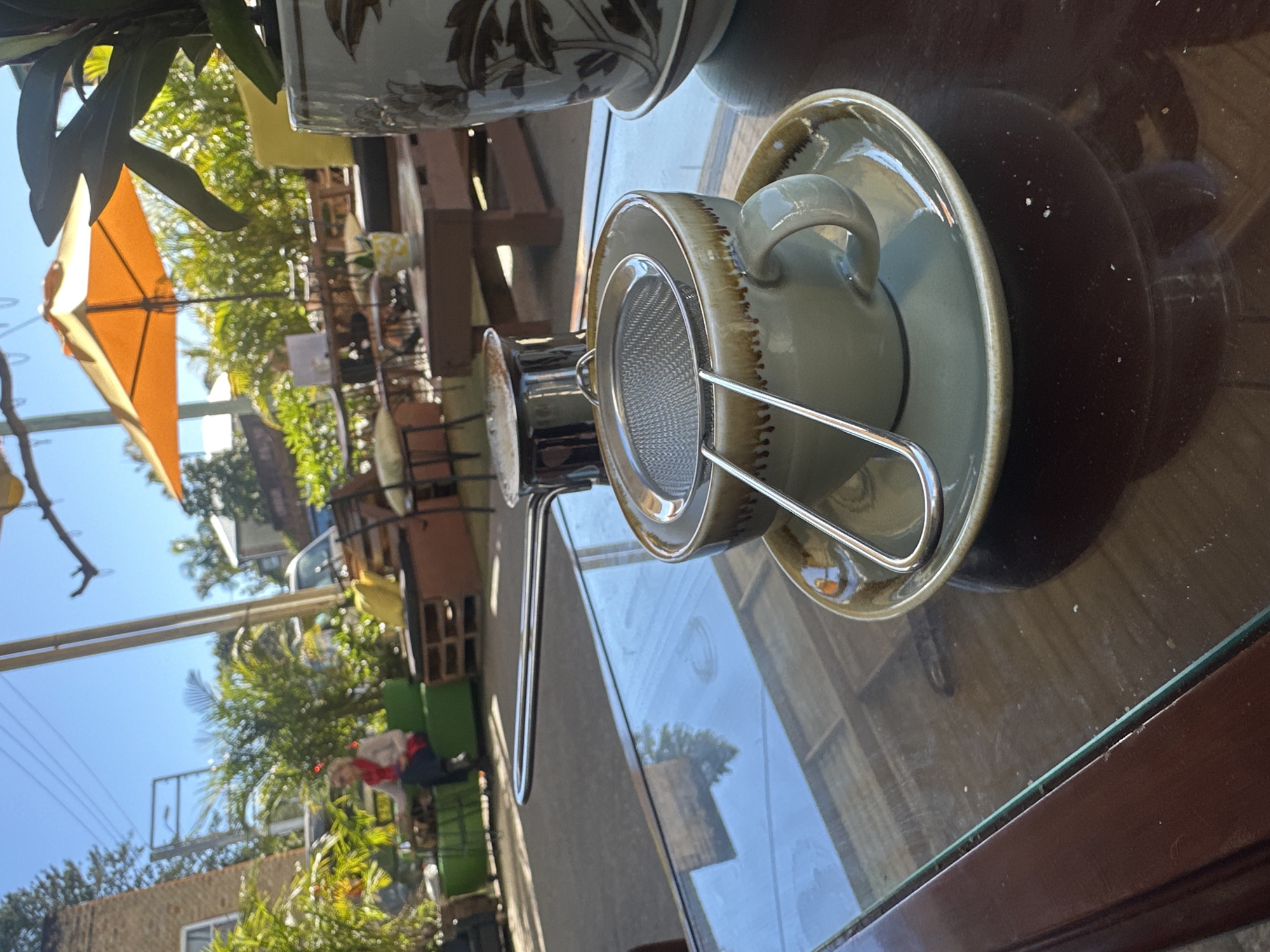 This image depicts an outdoor cafe scene in Bellingen, Australia, bathed in bright sunlight. In the foreground, a glass-topped table holds a ceramic coffee cup and saucer, with a metal tea strainer resting inside the cup. To the left of the cup, a small metal pot with a long handle is visible. The table's surface reflects the surrounding environment, including trees and sky. In the background, there is outdoor seating made of pallets and cushions, an orange umbrella, and lush green plants. A woman in a red scarf is seated on a green sofa in the middle ground. The overall setting suggests a relaxed cafe atmosphere.