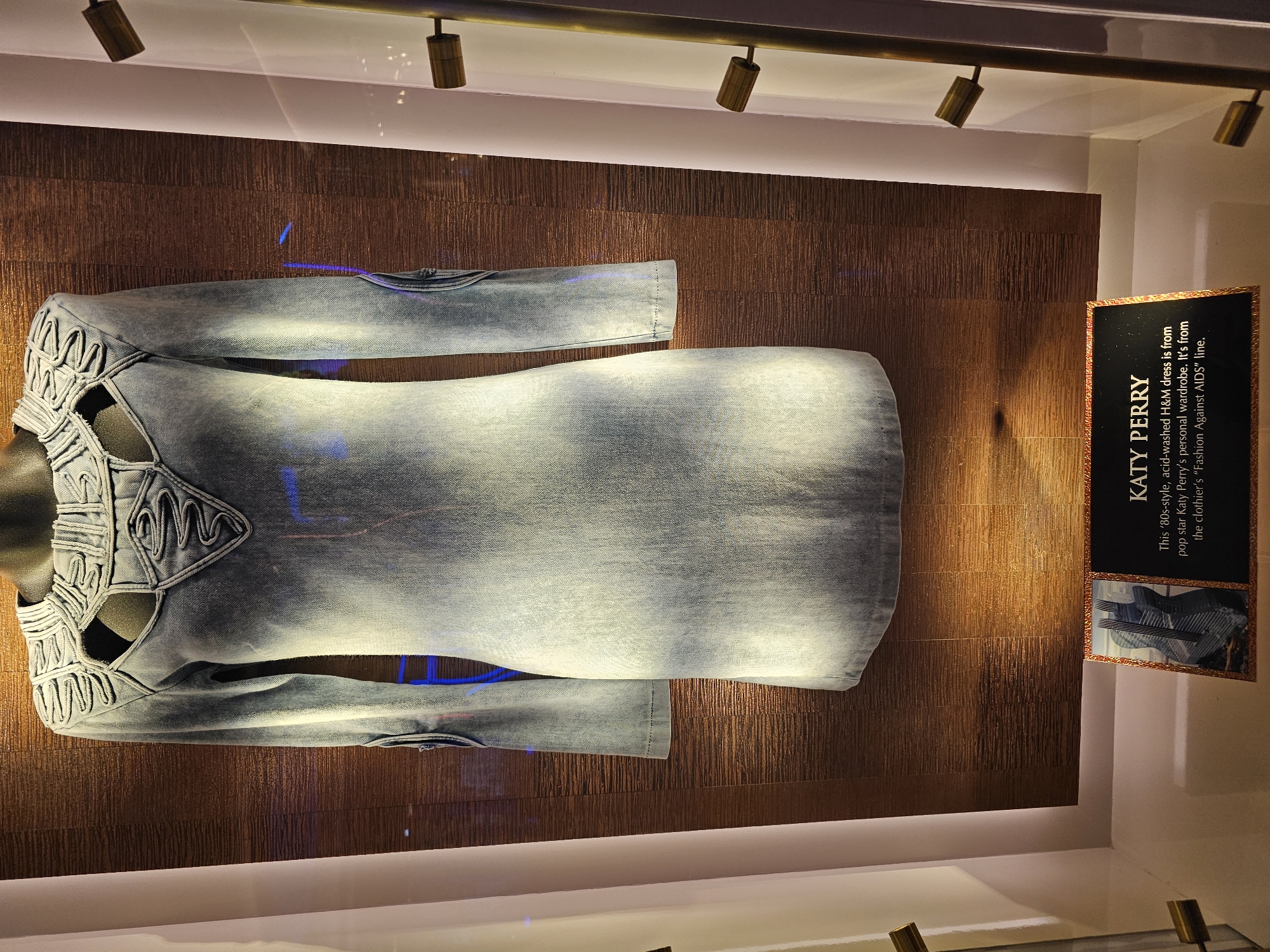 The image displays a dress, presumably showcased in a museum or exhibit, located in Hollywood Seminole Reservation, United States of America. The dress, made of acid-washed denim, features a form-fitting design with unique cutout details along the neckline and sleeves. It is mounted on a mannequin and positioned against a wall with a textured, bronze-colored finish. To the right of the dress, a placard bearing the title "KATY PERRY" provides information about the garment. The text on the placard states that the dress is from Katy Perry's personal wardrobe and is part of H&M's "Fashion Against AIDS" line. The scene is illuminated by overhead spotlights, casting shadows and highlighting the textures of the dress and the wall.