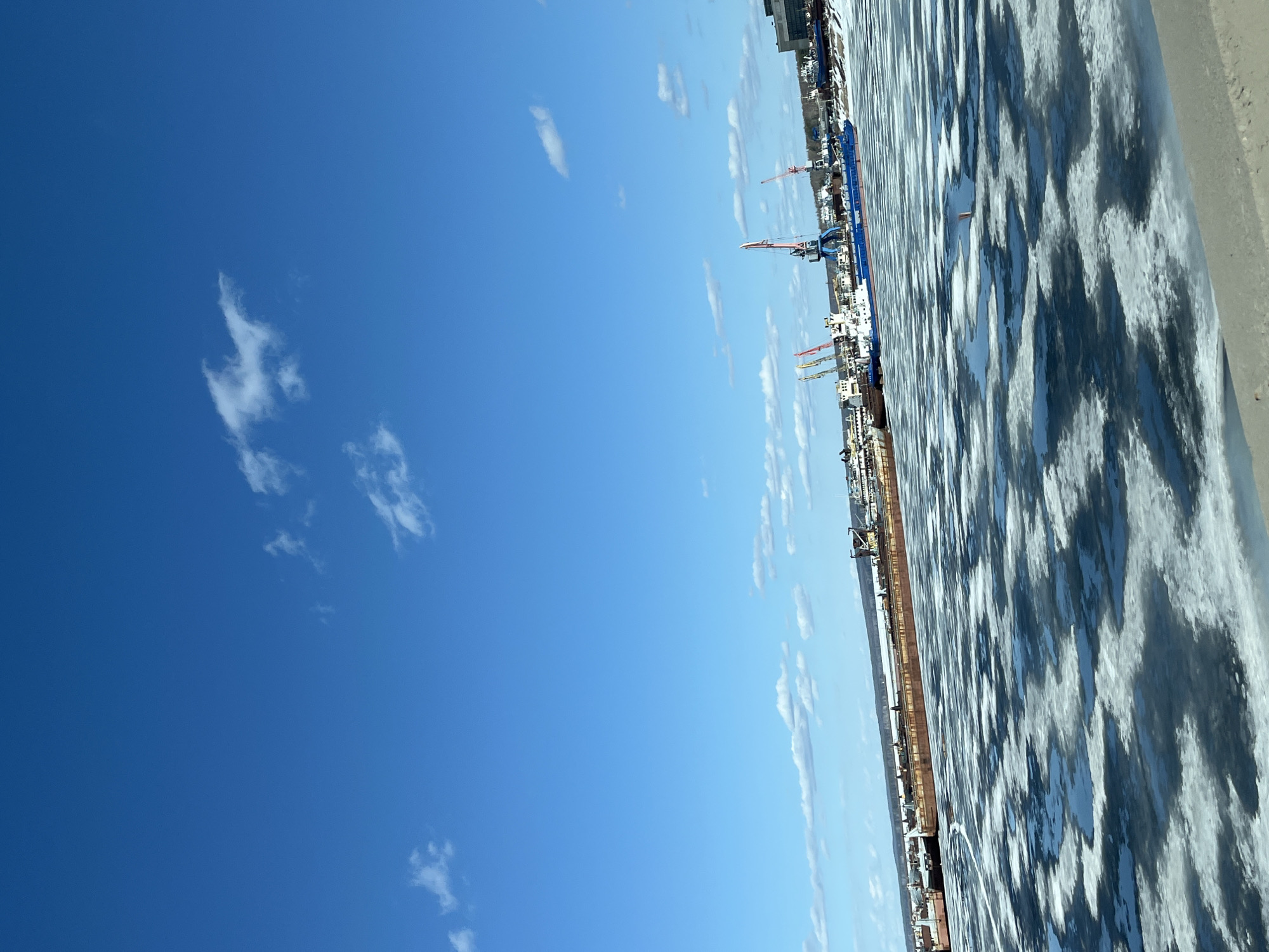 The image depicts the city of Chaikovsky, Russia, on a bright day with a clear blue sky dotted with scattered white clouds. The right side of the image shows a partially frozen body of water, with patterns of ice and water visible. A long cargo ship is docked along the shoreline, with several cranes and buildings visible in the background. The overall impression is that of a port or industrial area on a cold but sunny day.
