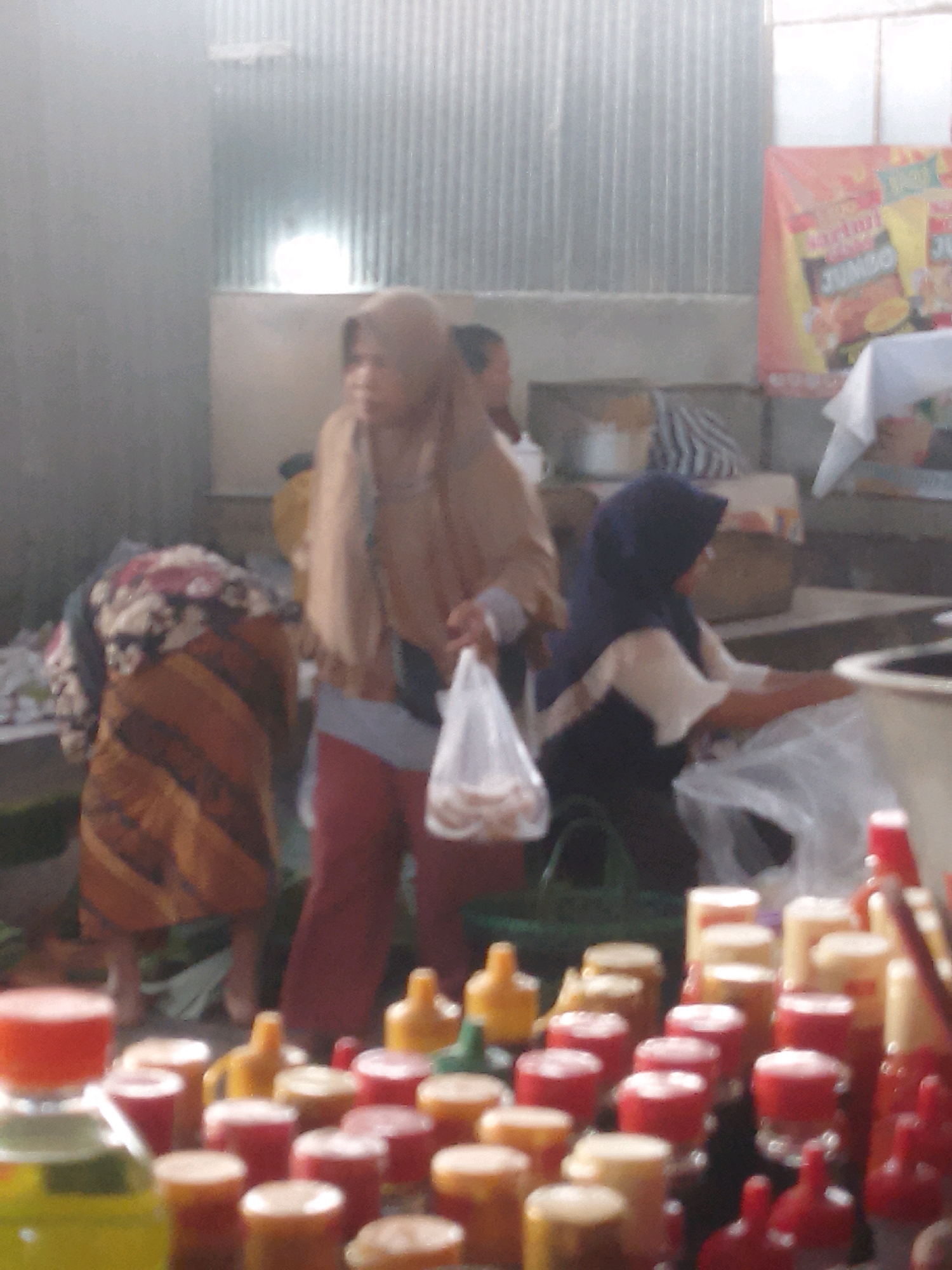 The image captures a street scene in Gondangrejo, Indonesia, likely a market or commercial area. In the foreground, an assortment of colorful bottles, some red and yellow, are arranged, suggesting they might contain liquids like sauces or drinks. In the mid-ground, three individuals are visible. The person on the left wears a patterned cloth and appears to be either bending down or looking downwards. The central figure, a woman wearing a light brown hijab and dark pants, is holding a clear plastic bag containing items. To the right, another individual in a dark blue hijab and a white top is interacting with something on a table, partially obscured by plastic sheeting. The background features a corrugated metal wall and a banner with text and graphics, indicating a commercial establishment. The overall impression is one of a busy marketplace, with people engaged in shopping or selling activities.