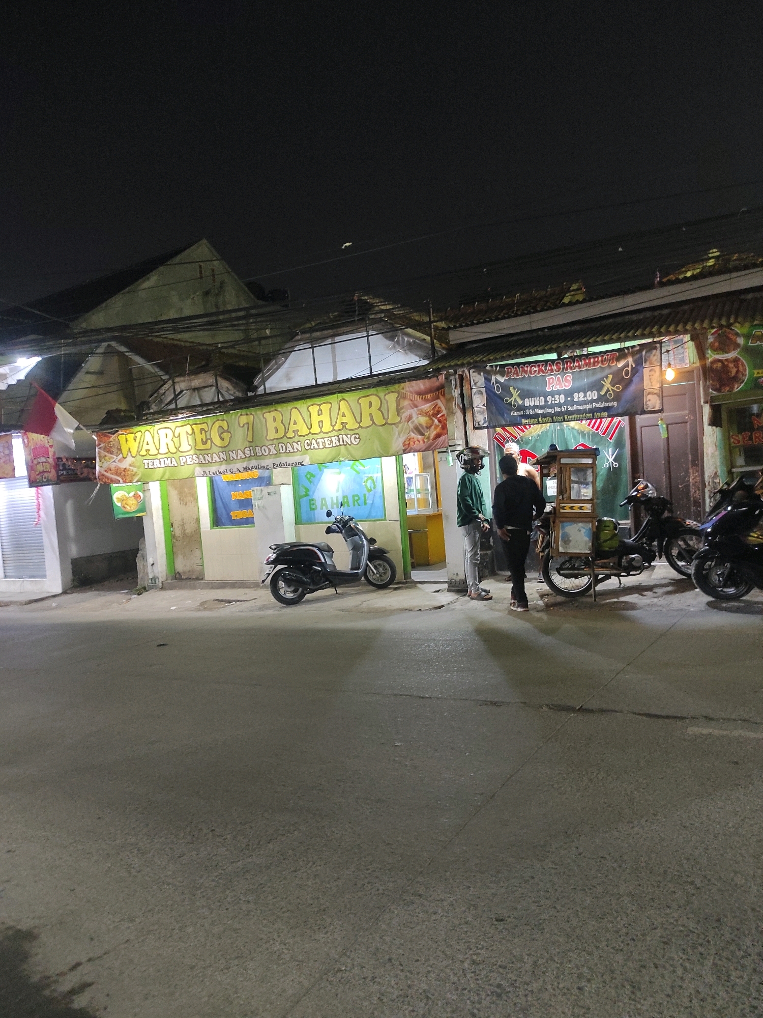 The image captures a street scene at night in Padalarang, Indonesia. A building with a green awning labeled "WARTEG 7 BAHARI TERIMA PESANAN NASI BOX DAN CATERING" is prominently featured. To the right, a banner for "PANGKAS RAMBUT PAS" (hair cutting salon) is visible, displaying operating hours from 9:30 to 22:00 and an address. Two men are standing near a food cart which appears to be part of the "WARTEG 7 BAHARI" establishment. Several motorcycles are parked along the street, including a black scooter in front of the warteg and other motorcycles further down. The environment is dimly lit, suggesting it is late at night. The street is paved, and overhead electrical wires are present.