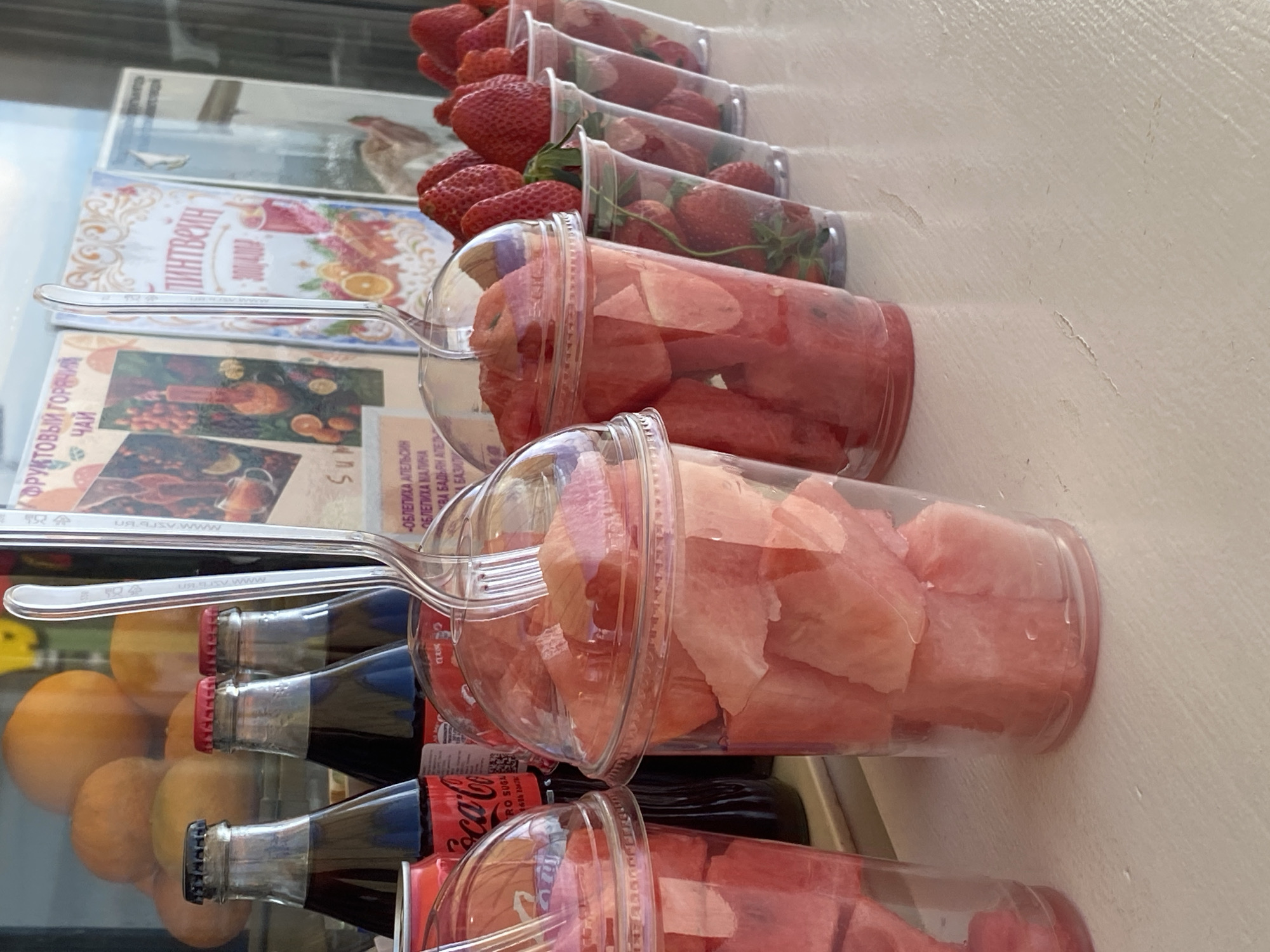 The image depicts a display of prepared fruits and drinks, likely in Sochi, Russia. There are rows of plastic cups filled with cut watermelon, and rows of cups filled with strawberries, all with plastic spoons inserted. Below the fruit are glass bottles of Coca-Cola and oranges. Behind the display are what appear to be books or printed materials with text in Cyrillic.