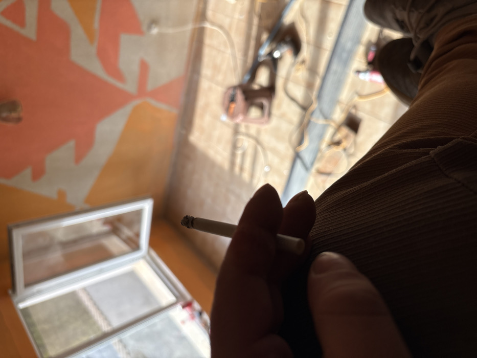 The scene depicts an interior room in the city of Stefan Karadja, Bulgaria, from a first-person perspective looking down and forward. In the immediate foreground, a right hand holds a lit, white cigarette, topped with dark gray ash. The individual is wearing light brown corduroy trousers and dark brown shoes or boots, partially visible at the bottom right.
The room exhibits signs of ongoing work or renovation. The walls are painted with an abstract, geometric pattern utilizing shades of orange and a lighter, possibly peach or beige, color. A white, double-pane window is open outward on the left, revealing a blurry outdoor area. The floor is tiled in a light, possibly beige or brown, color and is covered with multiple electrical cords, including a prominent yellow coiled cord. A brown industrial-style vacuum cleaner or similar equipment is positioned on the floor in the mid-ground, along with other dark objects, possibly tools. A small red object, appearing to be a canister or bottle, is visible near the bottom right. Sunlight illuminates the floor, casting distinct shadows. A small, dark fixture is mounted on the ceiling in the upper mid-ground.