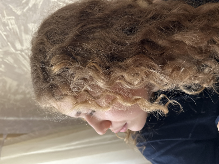 The image depicts a close-up, profile view of a single individual within an indoor setting in Nizhnekamsk, Russia. The subject, a person with voluminous, light brown to blonde curly hair, is positioned facing left, with their gaze directed slightly upward and to the left of the frame. Visible facial features include a prominent nose, partially visible lips, and the contour of the cheek and forehead. The individual is wearing a dark-colored (likely navy blue or black) shirt or top, partially visible on the left shoulder and upper arm.
The background consists of two distinct areas. To the left, a light-colored, possibly beige or white, curtain or fabric is visible, illuminated by soft light. To the right and behind the subject's head, a light-toned wall features a subtle, textured pattern, with faint, elongated shadow patterns cast upon it, consistent with light filtering through an external object like foliage. The scene is illuminated by natural light, primarily from the left, creating highlights on the subject's hair and face.