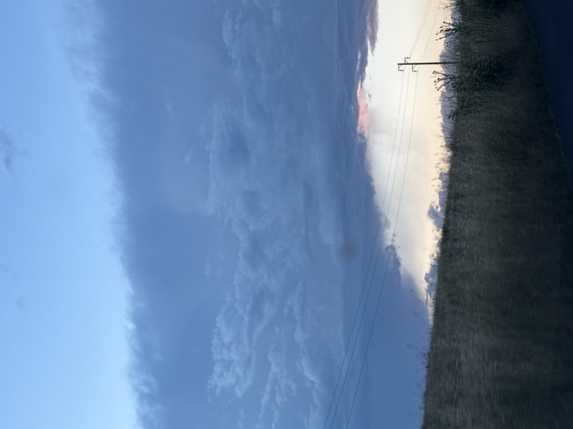 The image captures a rural landscape in the vicinity of Tvarditsa, Bulgaria, under a twilight sky. The foreground is dominated by a dry, grassy field that appears to be on a gentle slope. A dark, paved surface, possibly a road, is visible at the bottom right edge. In the middle ground, a utility pole with multiple wires stretches across the scene, connecting to unseen infrastructure. Beyond the field and wires, a soft, pastel-colored horizon suggests the fading light of day. The sky above is a gradient of blues, with heavy, dark clouds dominating the upper portion and lighter, wispy formations lower down. A hint of pinkish light is visible through a gap in the clouds near the horizon, indicating the last rays of sunlight. The overall atmosphere is serene and natural, devoid of any human figures or overt signs of activity.